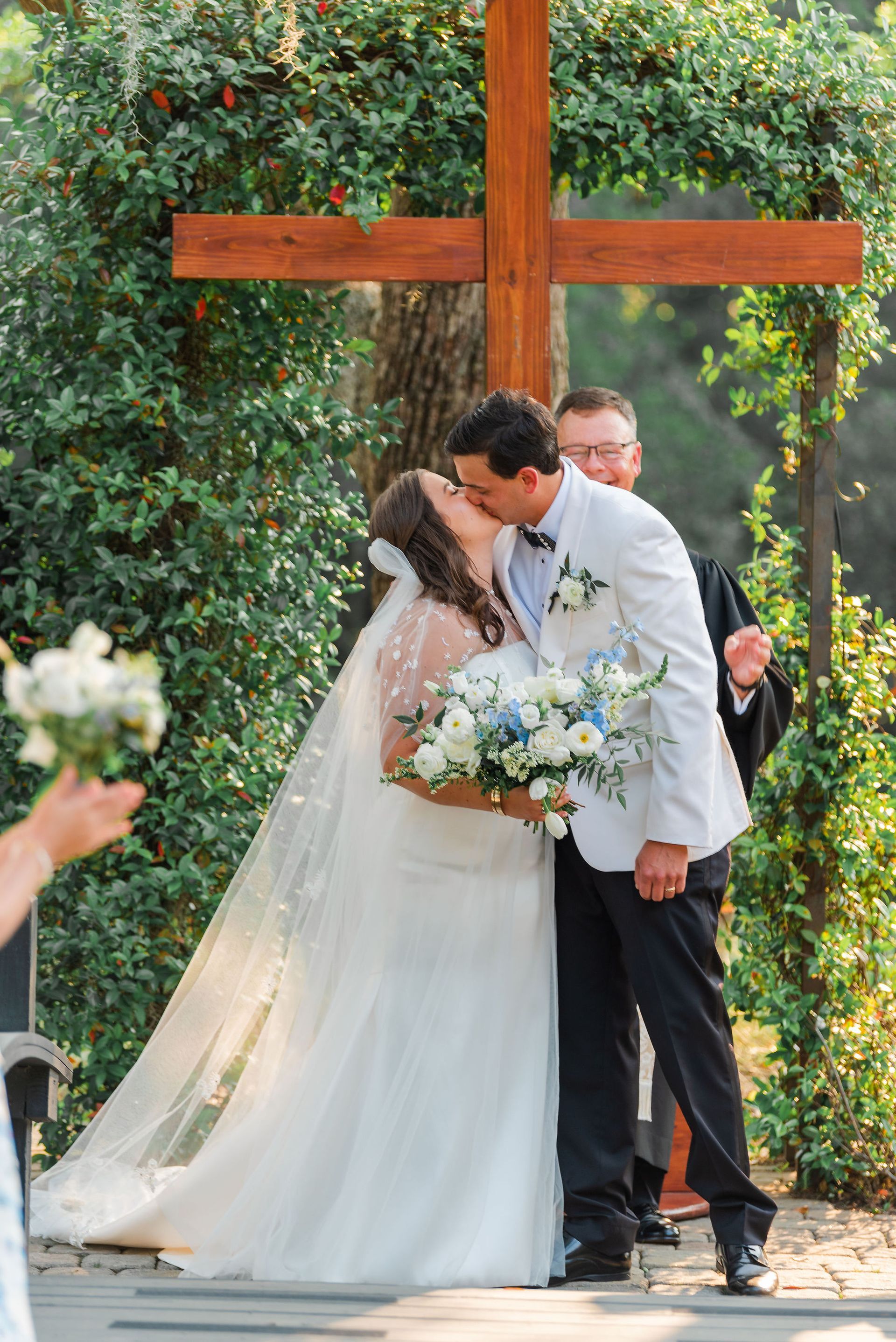 Bride and groom kiss under a wooden cross during an outdoor wedding ceremony