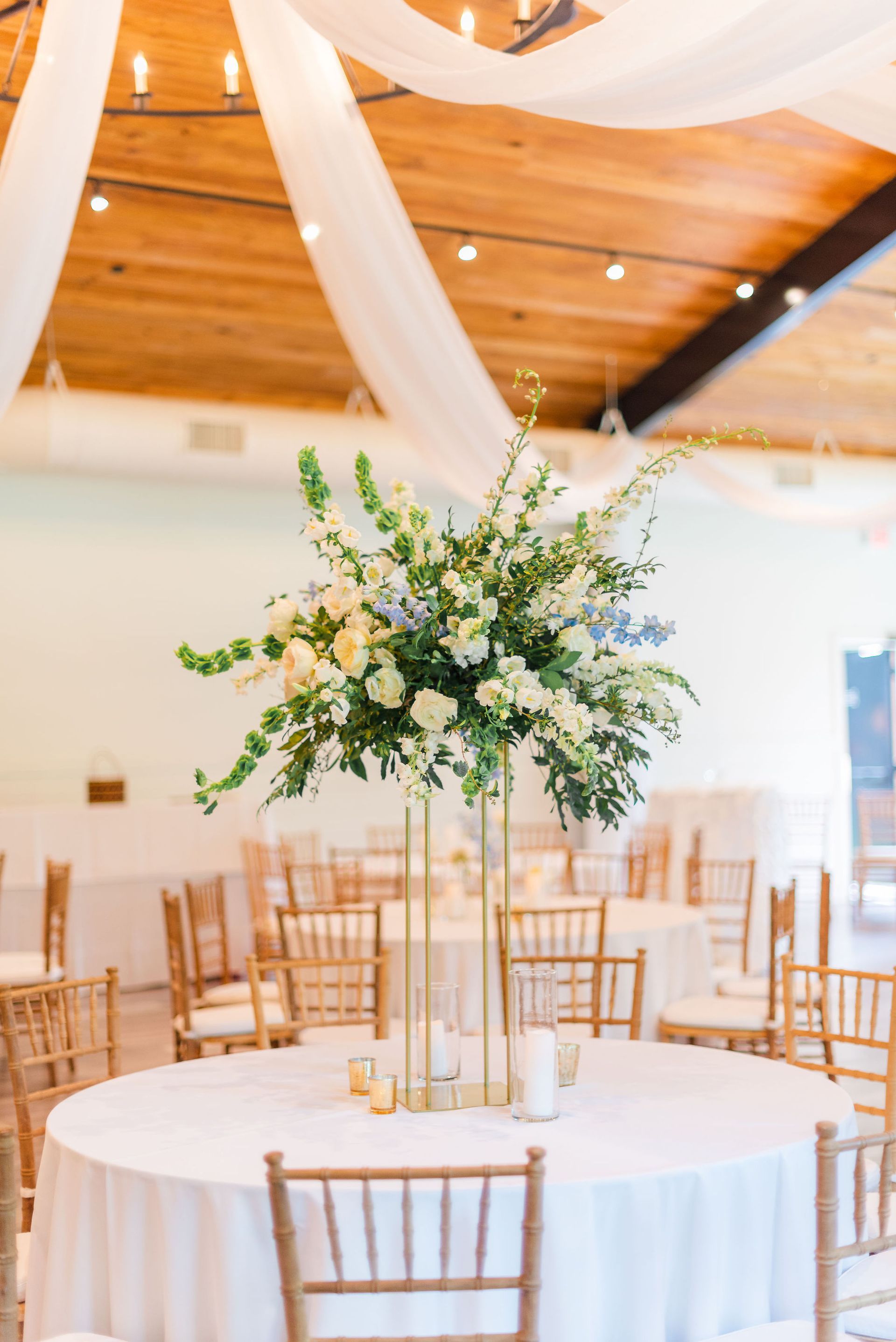 Elegant wedding reception table with white linens, gold chairs, and a tall floral centerpiece under a wooden ceiling