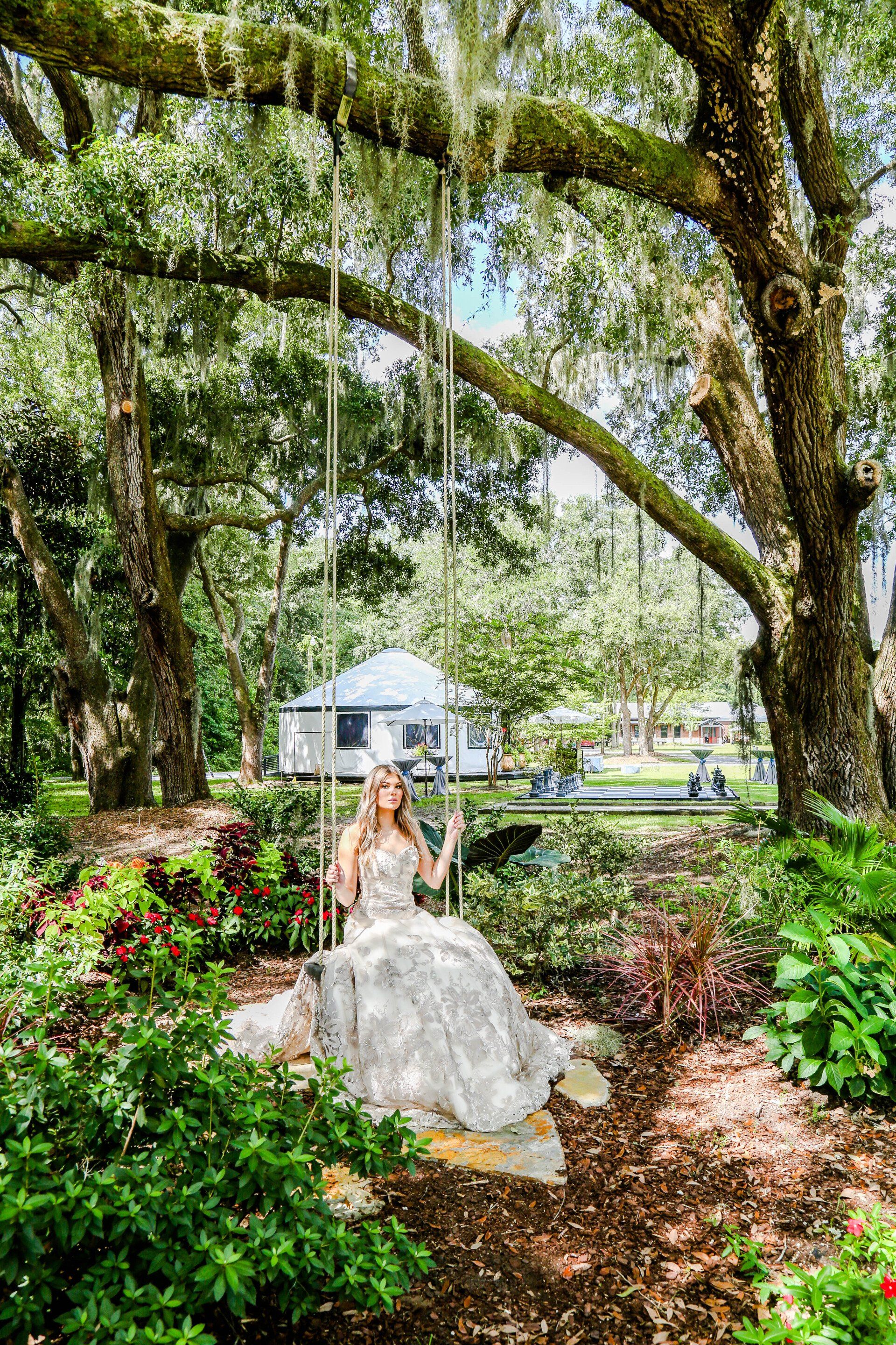 Bride standing on a rock in a lush garden under mossy oak trees, with a white tent in the background