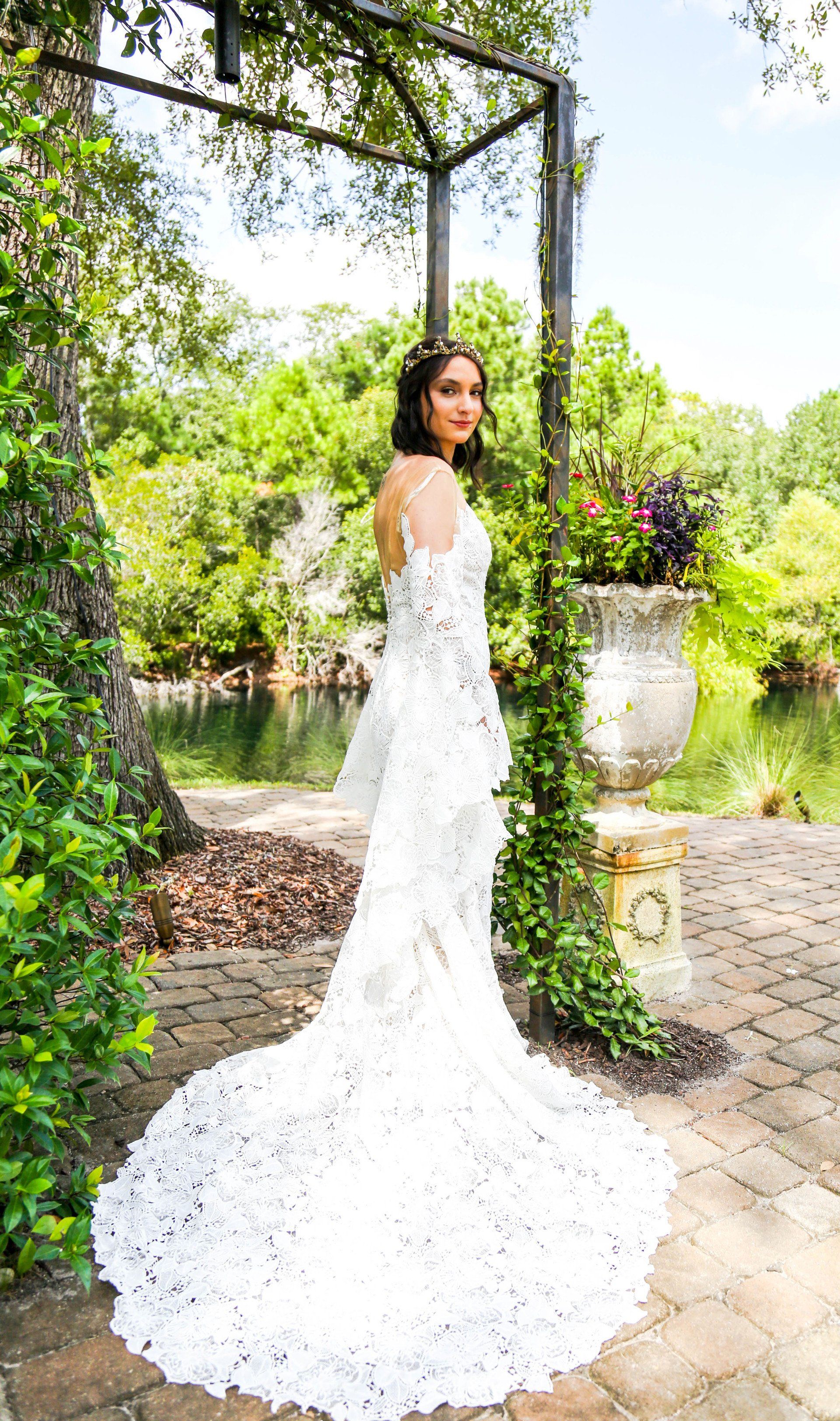 Woman in a long white lace gown standing in a sunlit garden by a vine-covered arch.