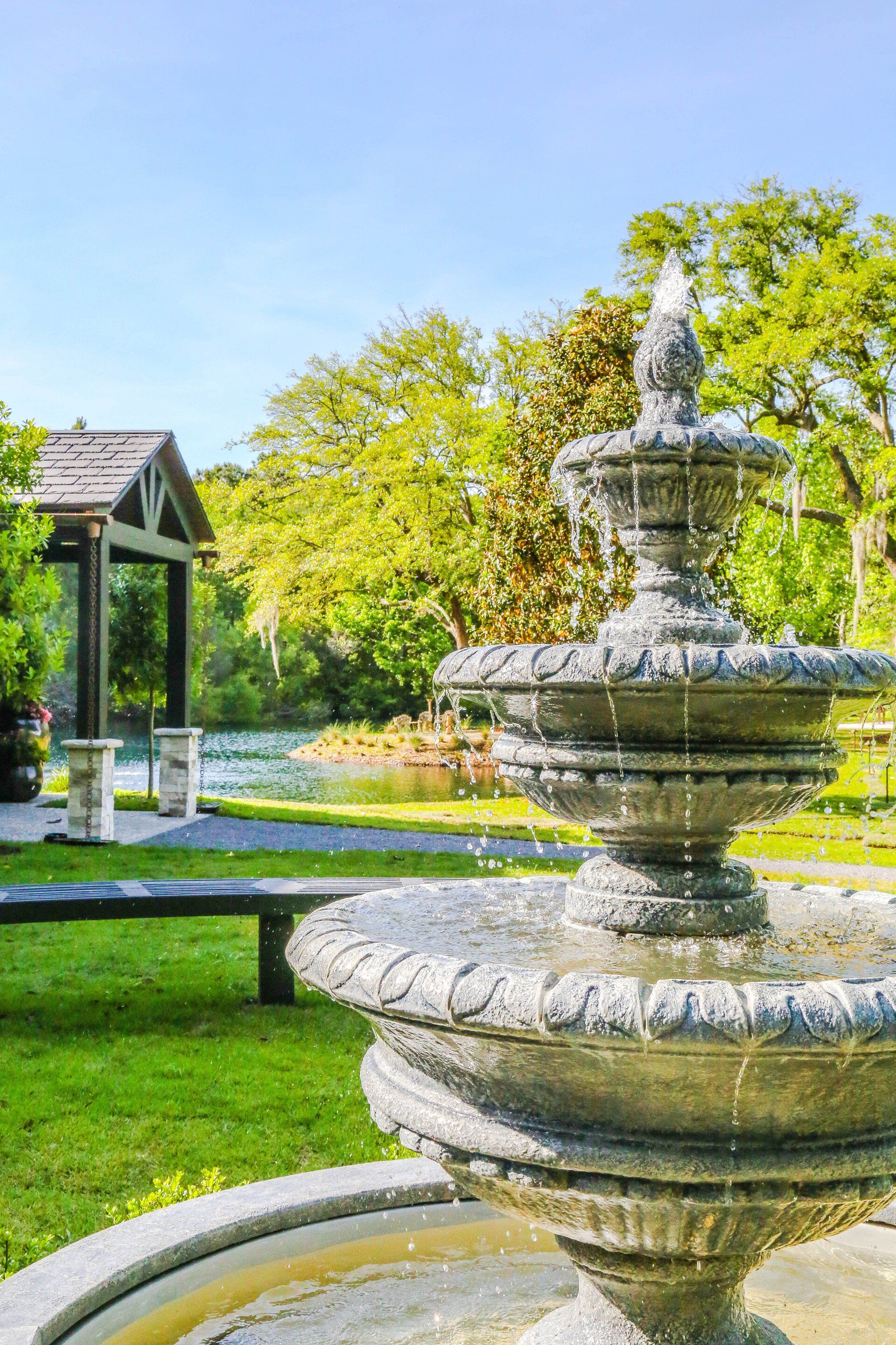 Stone fountain in a sunny garden with green grass, trees, and a gazebo in the background