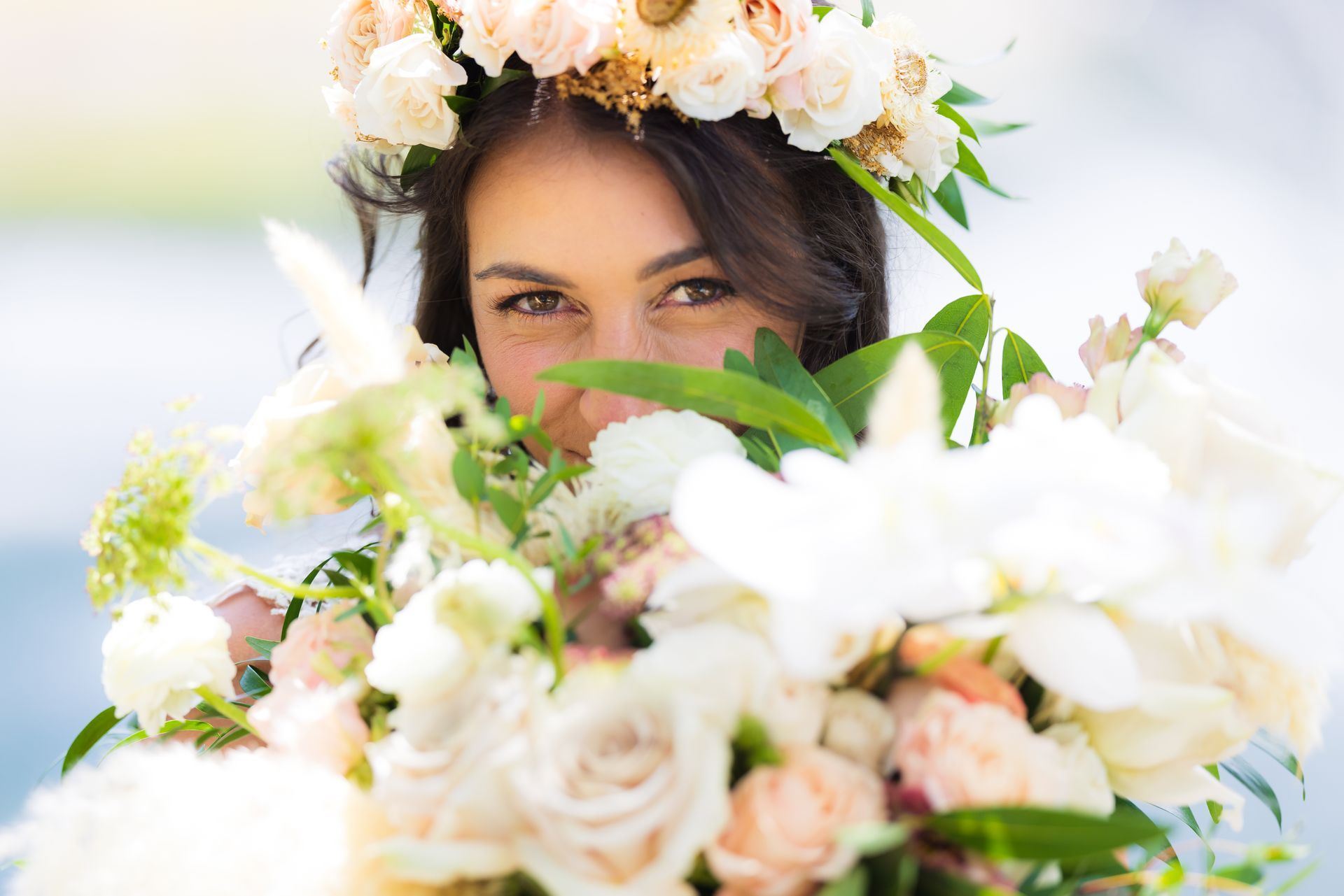 Woman framed by white and blush flowers, peeking through a lush floral arrangement outdoors.