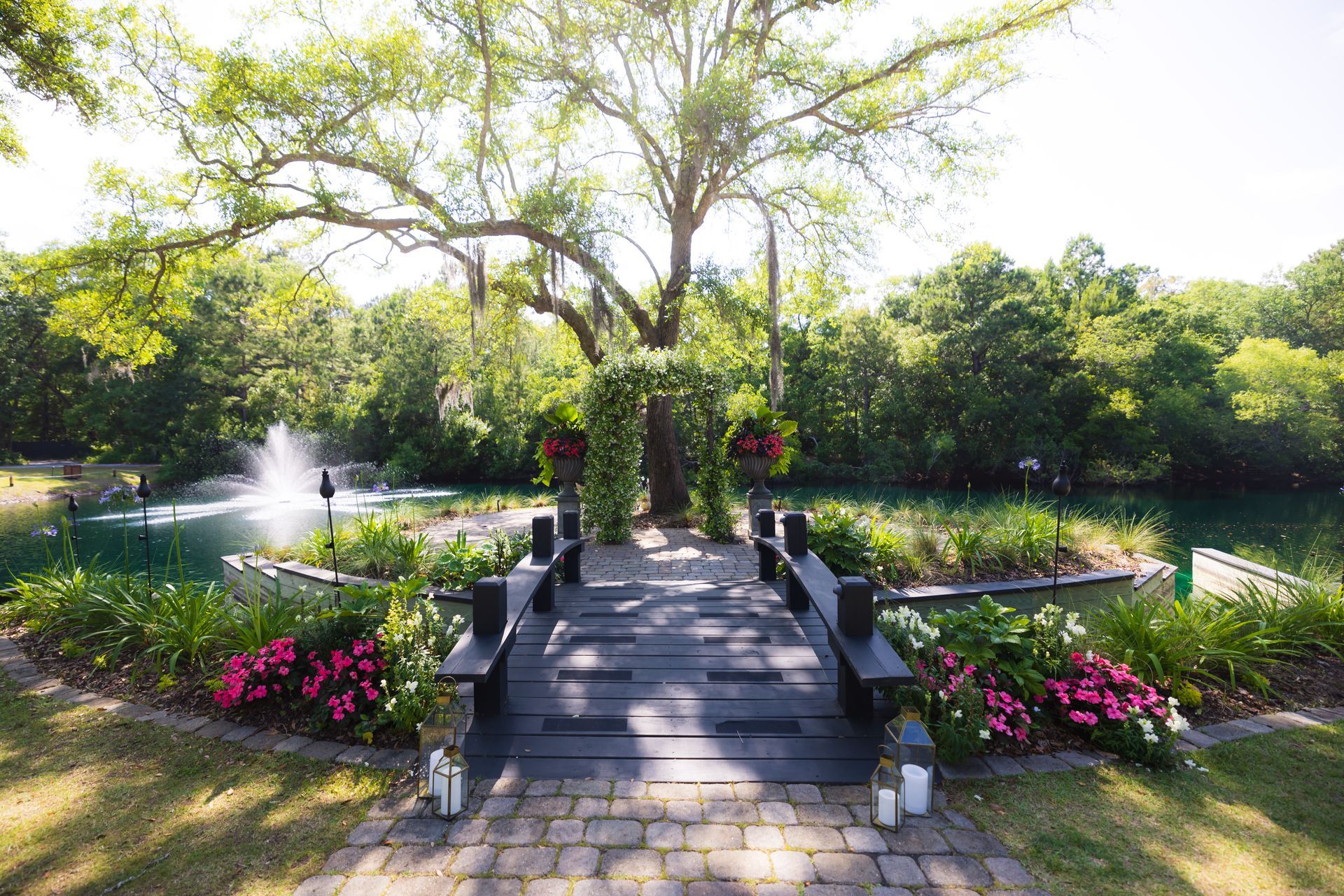 Stone bridge over a garden pond with fountains, pink flowers, and lush green trees in bright sunlight
