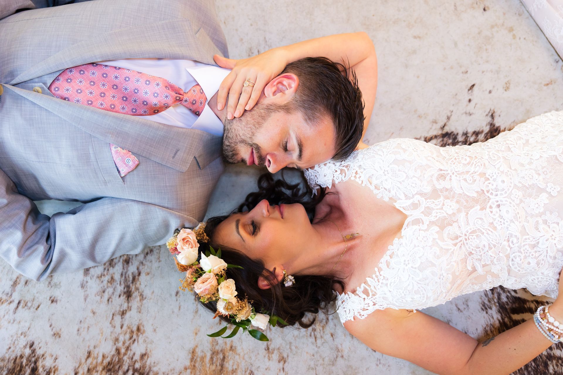 Couple in wedding attire lying together on a textured floor, touching foreheads and smiling softly