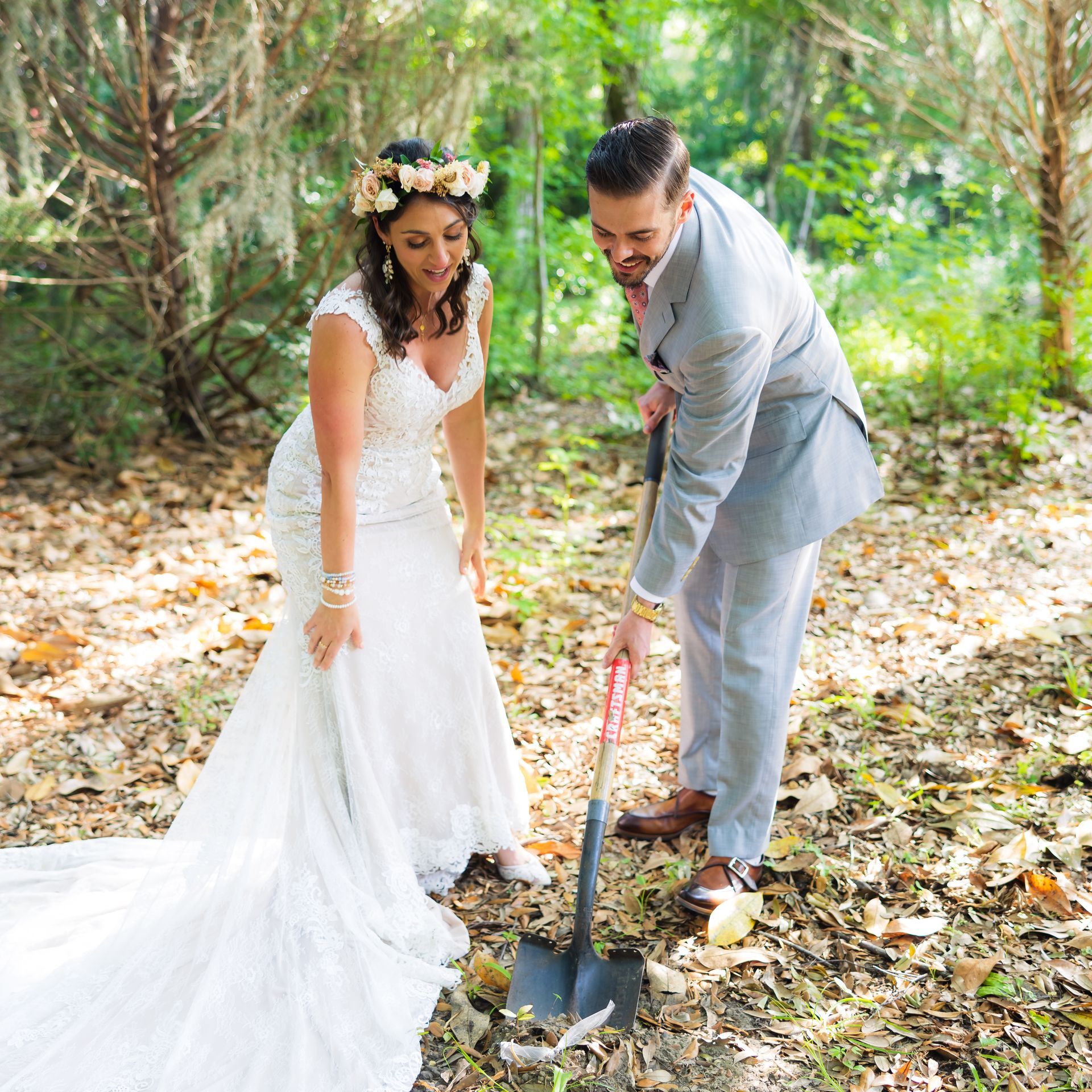 Bride and groom in a wooded setting, smiling while holding a shovel over a planted sapling