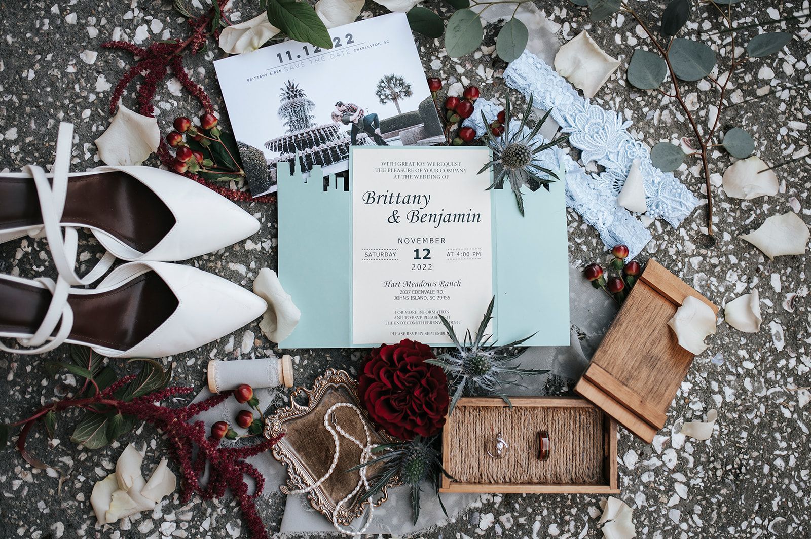Rustic flat lay of wedding stationery, white shoes, dried flowers, and a wooden ring box on a speckled stone background