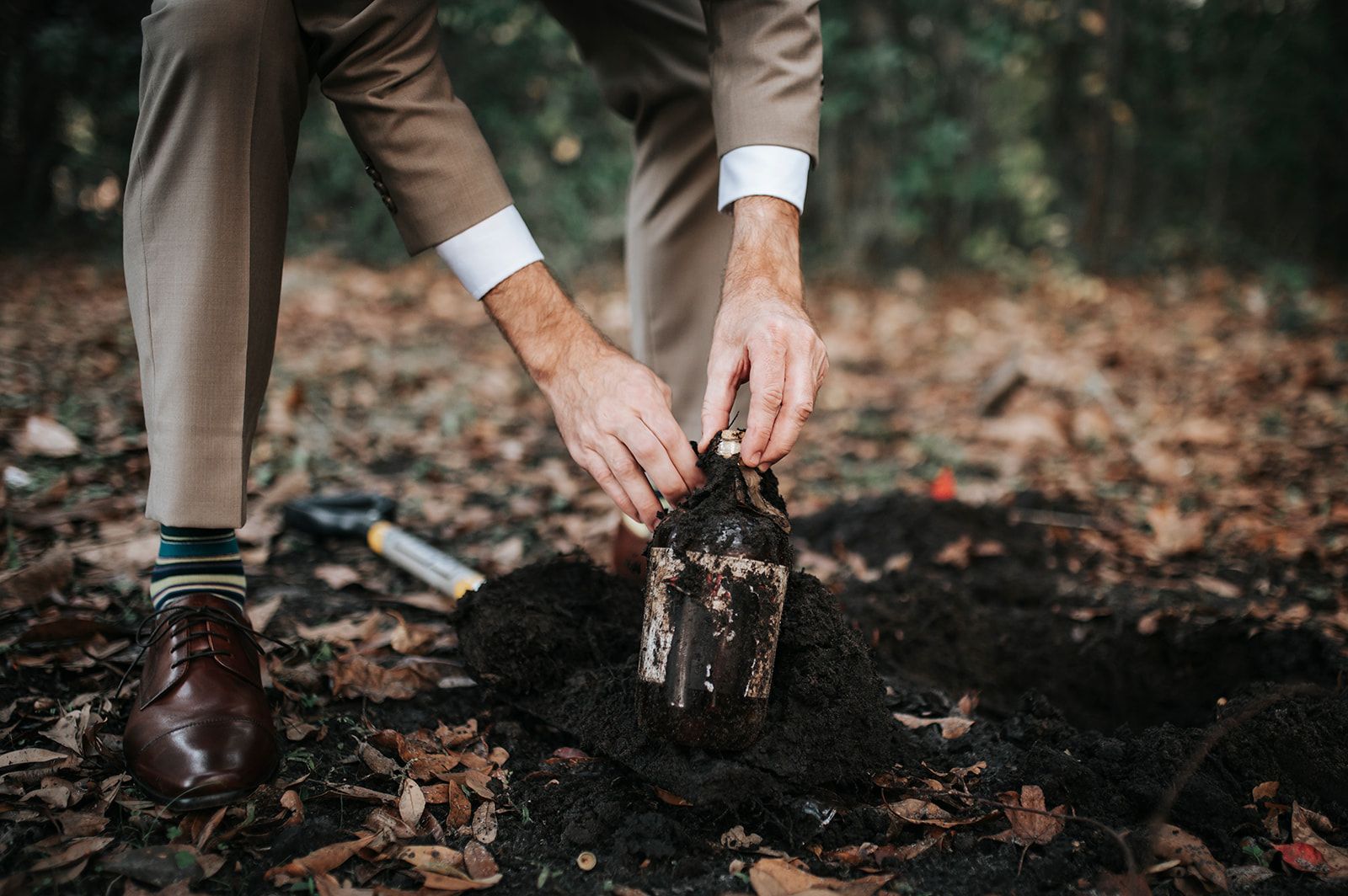 Hands planting a small sapling in dark soil outdoors, with a trowel nearby.