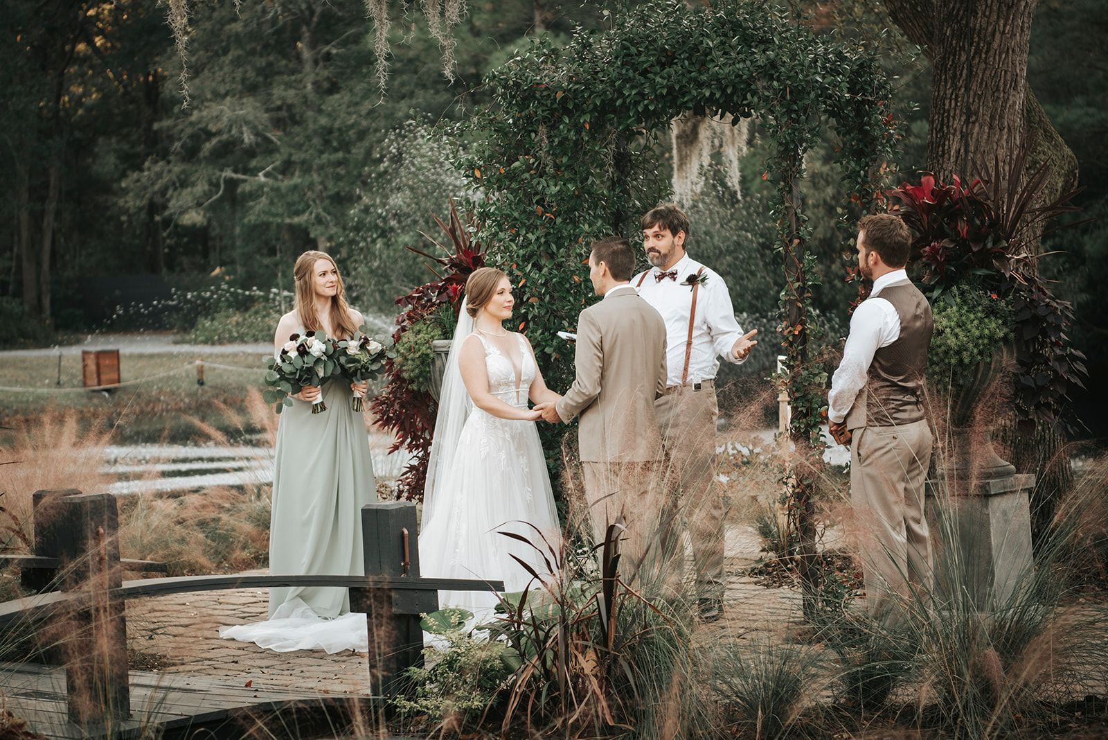 Wedding party standing by a pond in a wooded garden, holding bouquets and wearing formal attire.