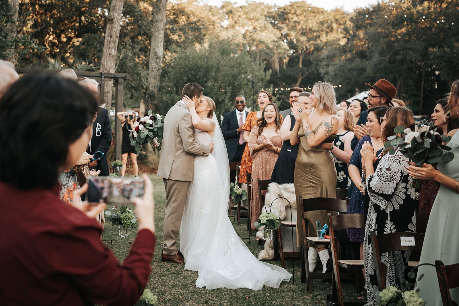 Outdoor wedding ceremony with bride and groom kissing, surrounded by applauding guests and autumn greenery