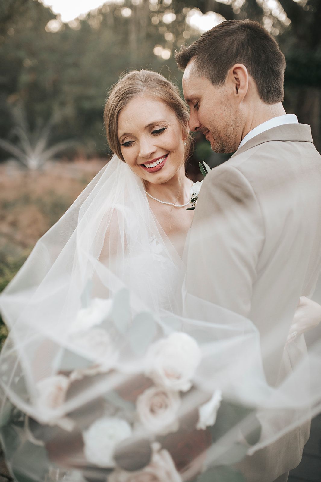Bride and groom smiling in a soft outdoor wedding portrait, veil flowing in the foreground