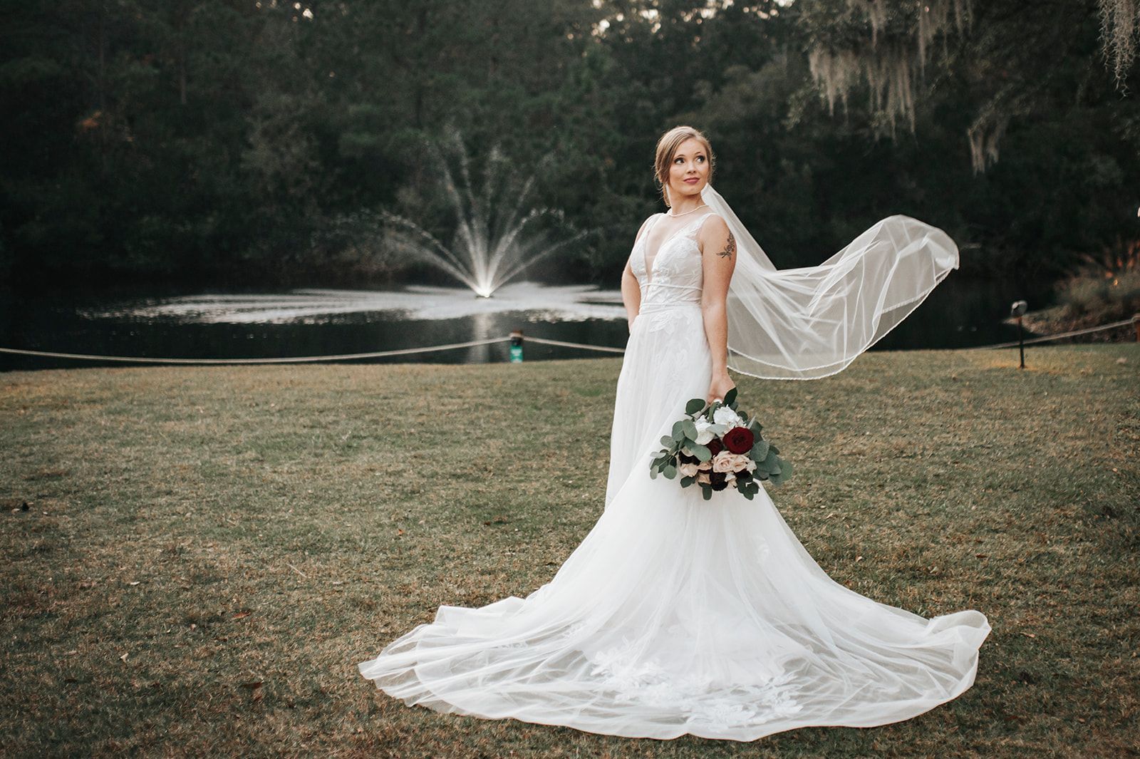 Bride in a white gown standing on grass beside a lake fountain, holding a bouquet with her veil flowing.