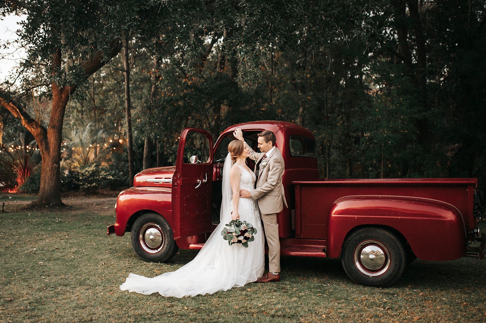 Bride and groom posing by a red vintage pickup truck in a wooded outdoor setting