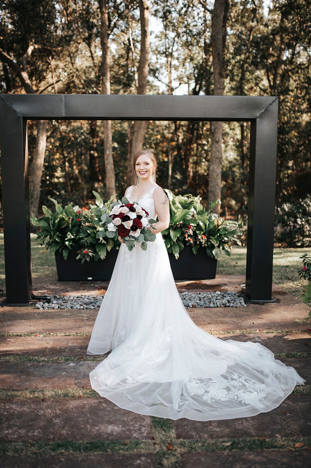 Bride in white gown holding bouquet beneath a dark arch in a wooded outdoor setting