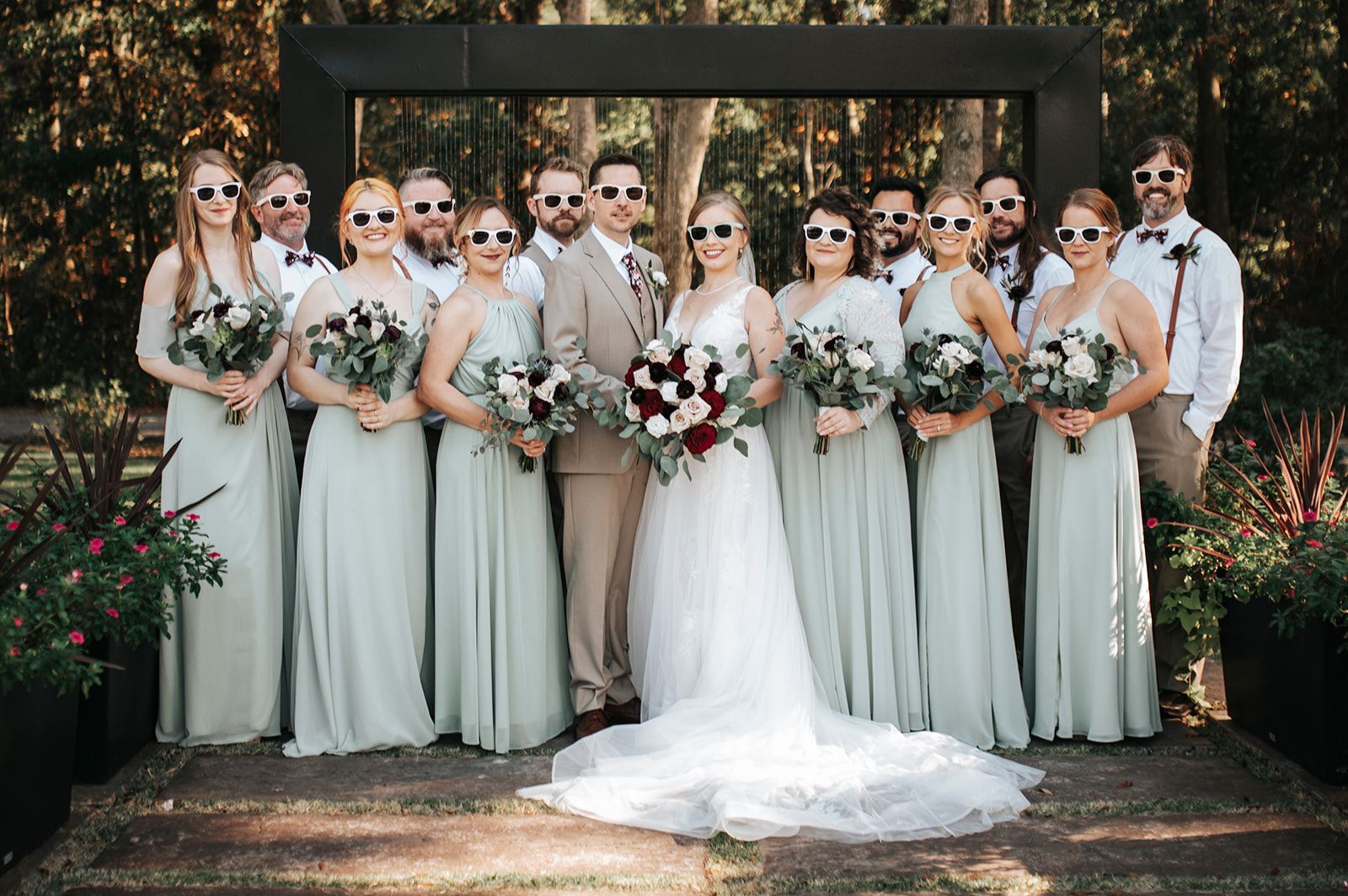 Wedding party posing outdoors under a dark arch, bridesmaids in sage dresses holding white bouquets