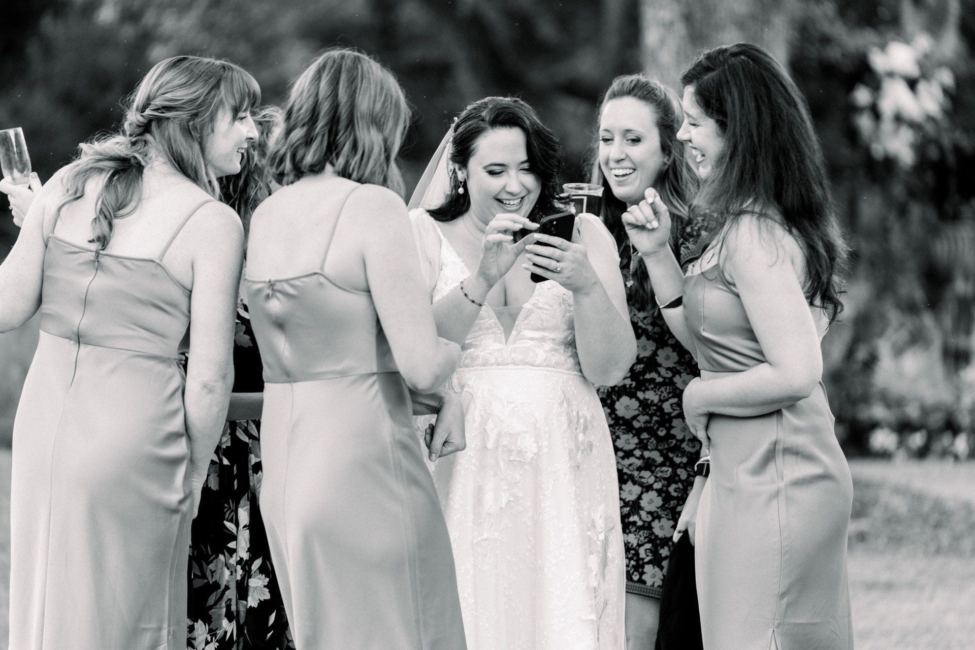 Group of women in dresses gathered around a smiling bride at an outdoor celebration, looking at a phone.