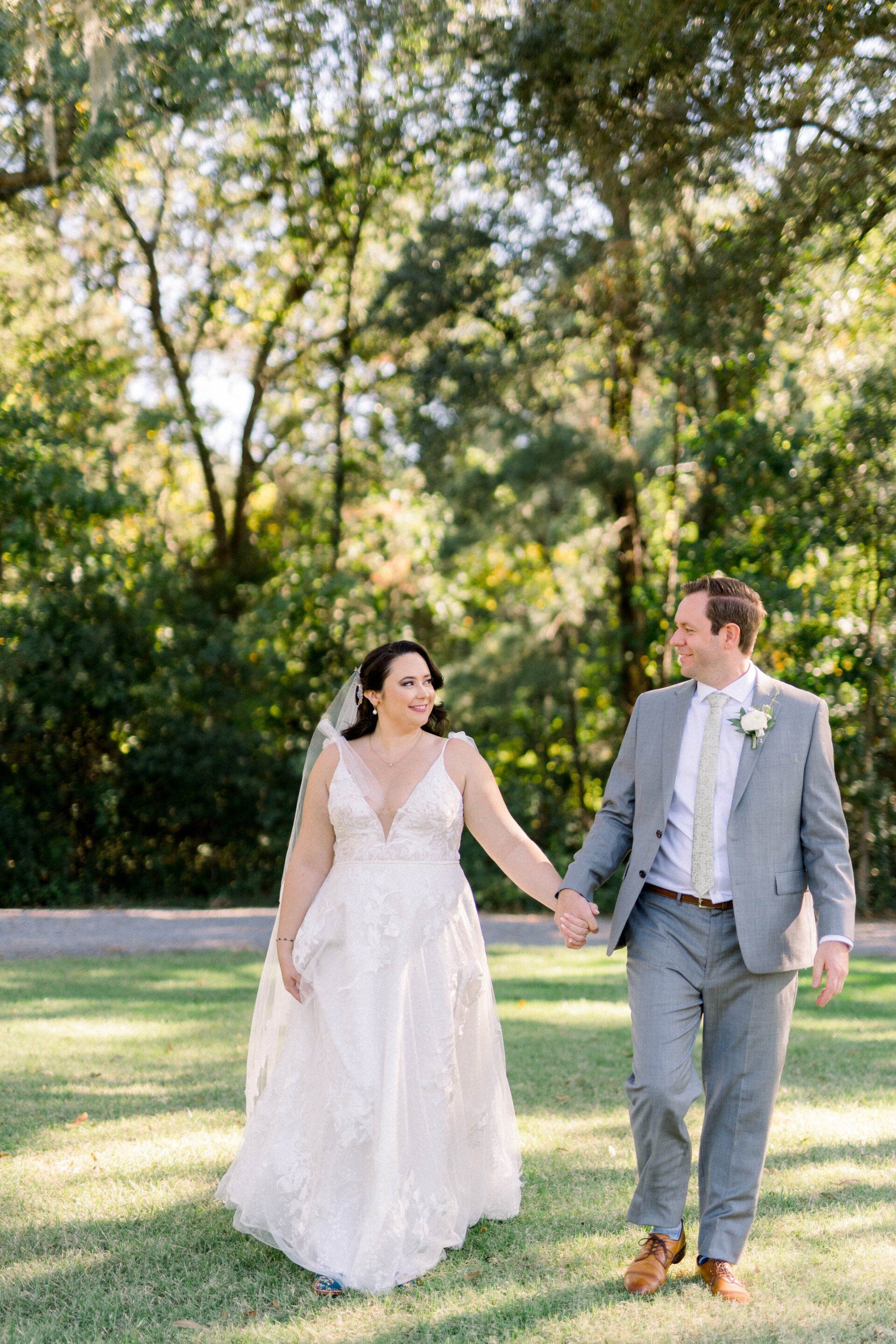 Couple in wedding attire holding hands while walking through a sunny park.