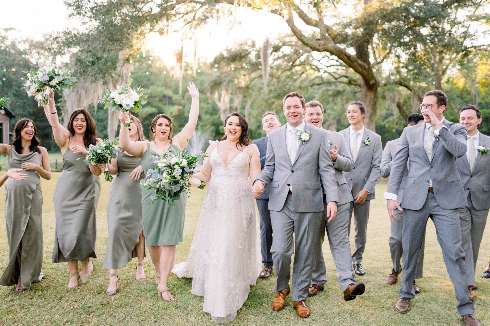 Wedding party walking on grass outdoors, bride in white and others in gray suits and dresses, holding bouquets.