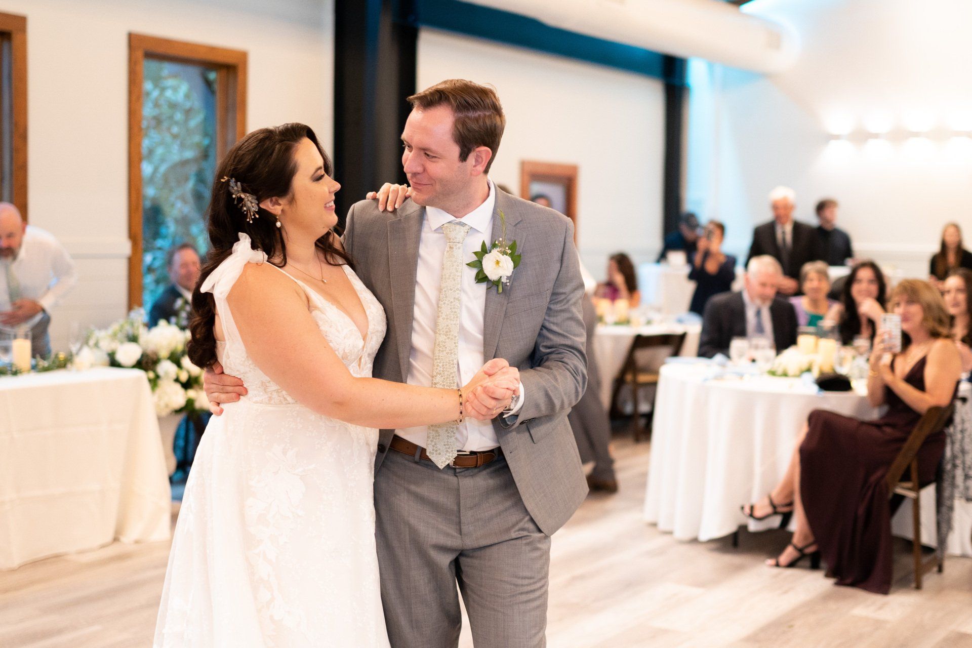 Couple dancing at a wedding reception in a bright banquet hall, guests seated at round tables behind them