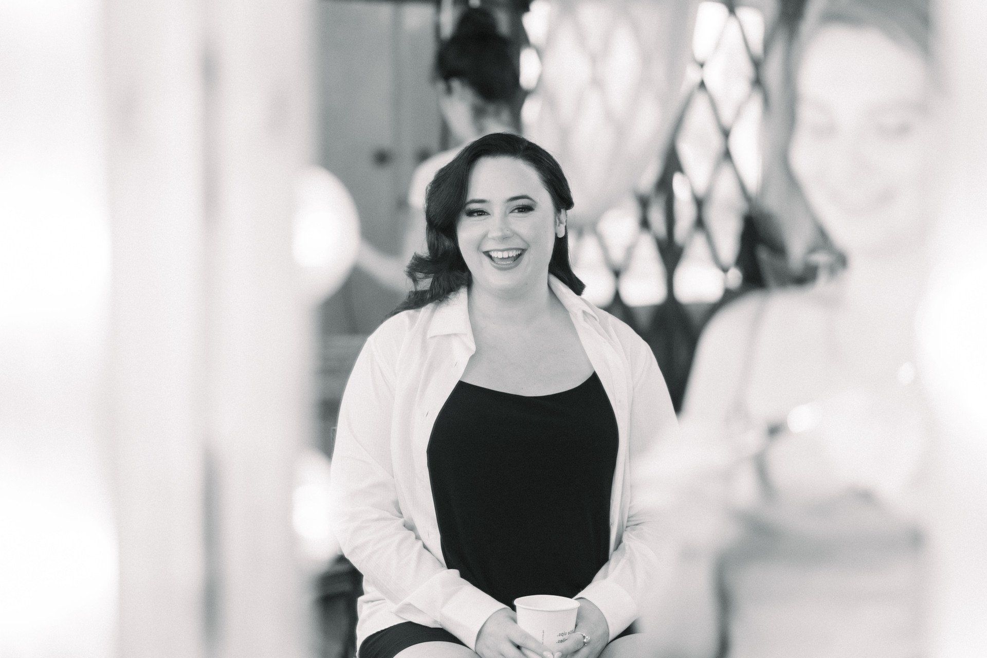 Smiling person seated indoors in a bright, softly blurred setting, wearing a white cardigan over a black top