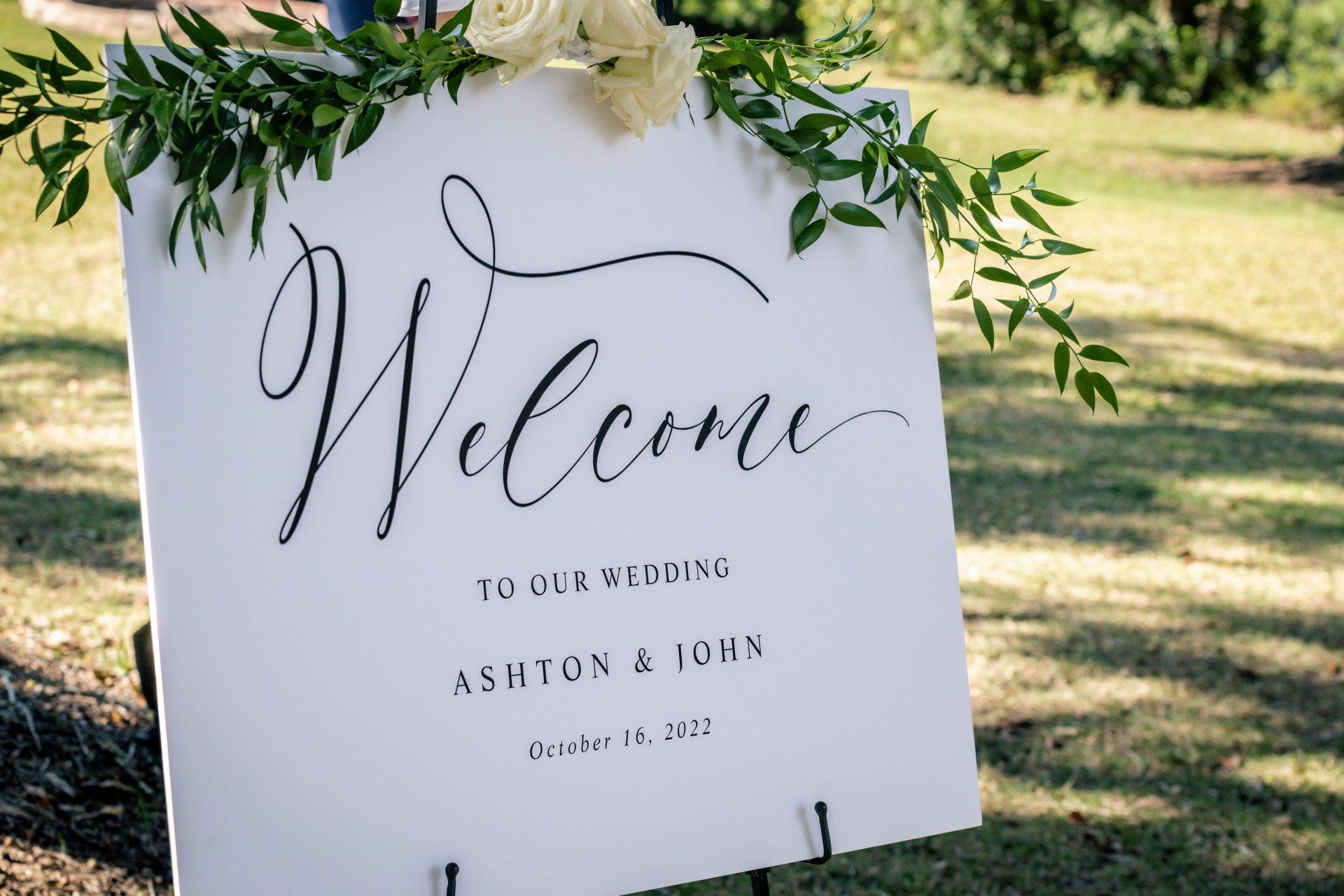 White welcome sign with greenery, set outdoors on grass.