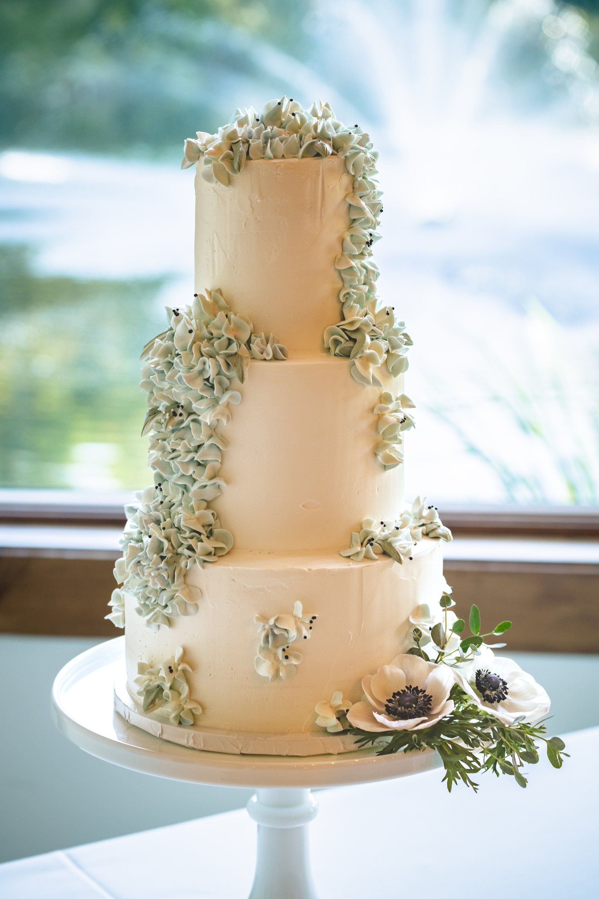 Three-tier white wedding cake with cascading white flowers on a white stand by a window