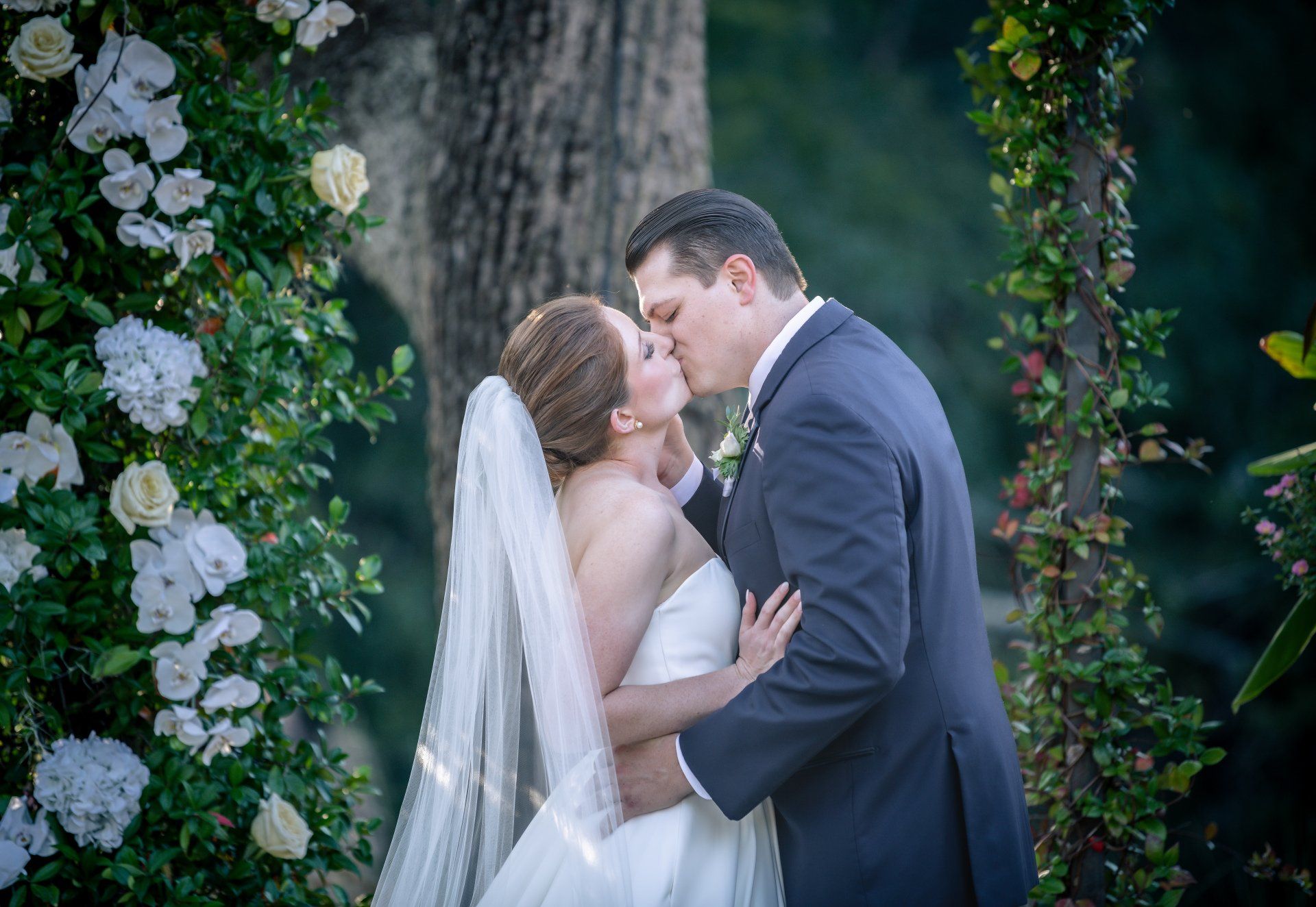Bride and groom kissing outdoors amid lush greenery and white flowers