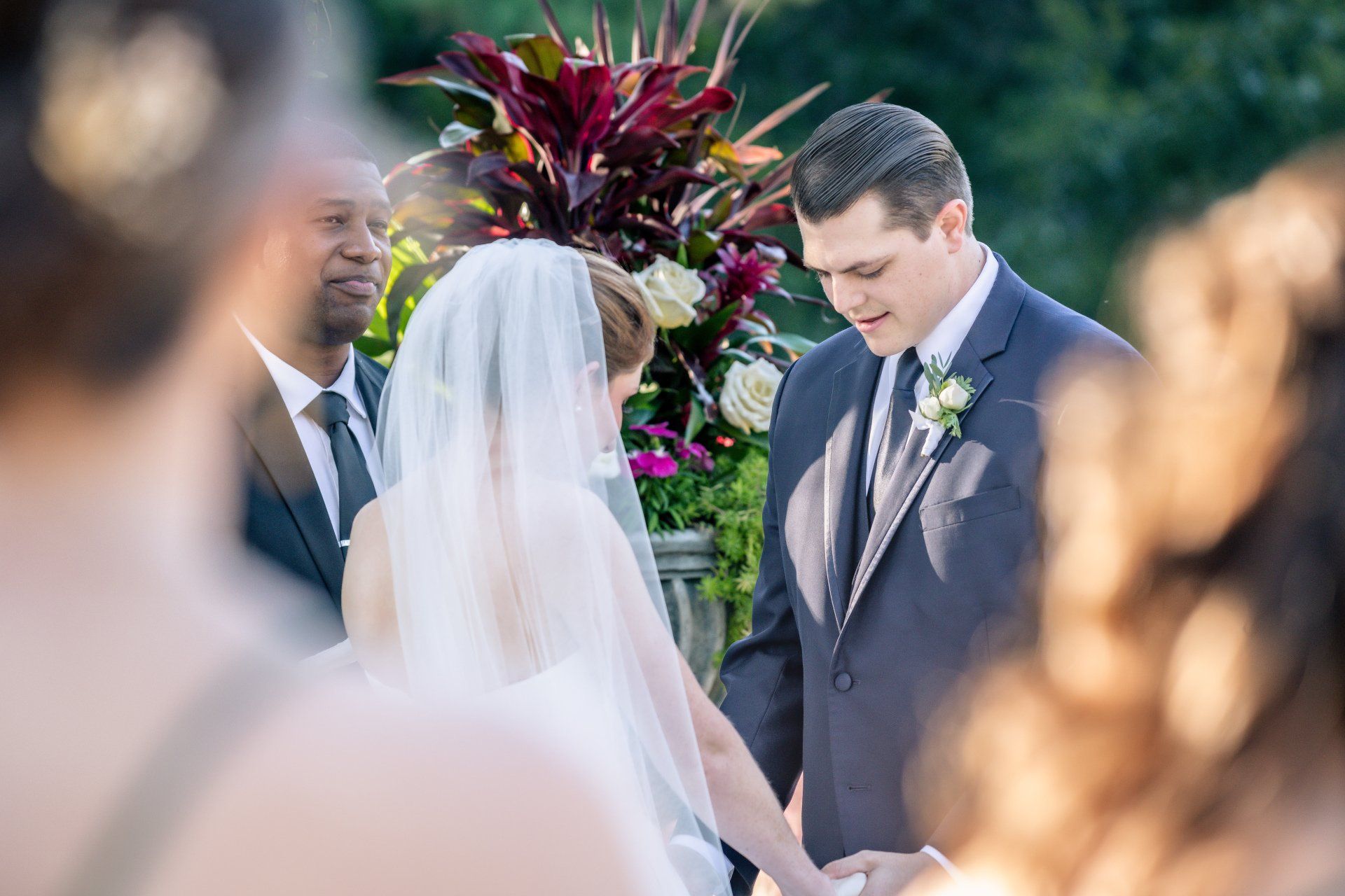 Couple exchanging vows outdoors before a floral arrangement, with the bride in a veil and the groom in a gray suit