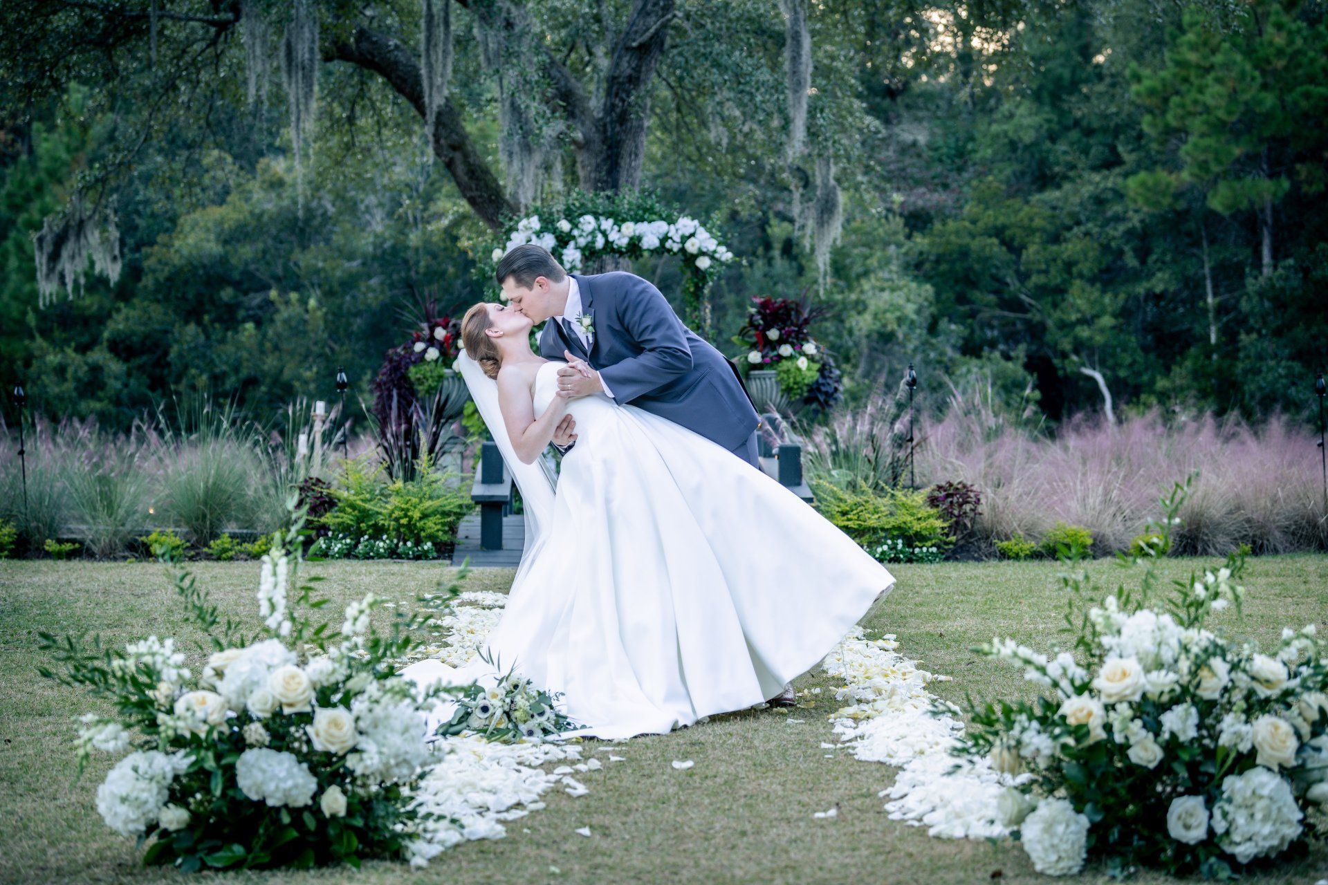 Bride and groom kissing in a garden wedding ceremony, framed by white flowers and greenery.