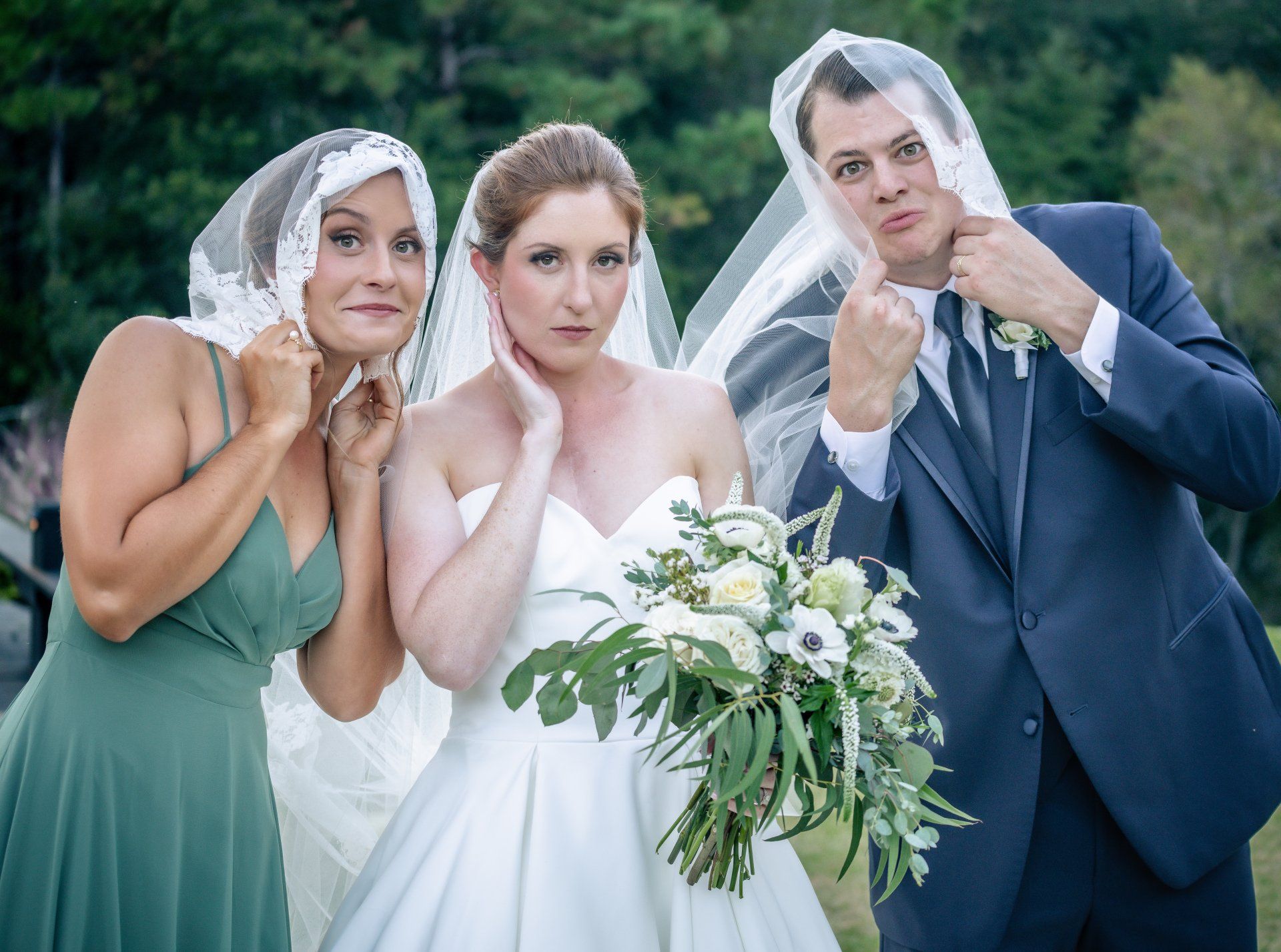 Three wedding guests in formal attire hold sheer veils over their heads outdoors, posing by a bride.