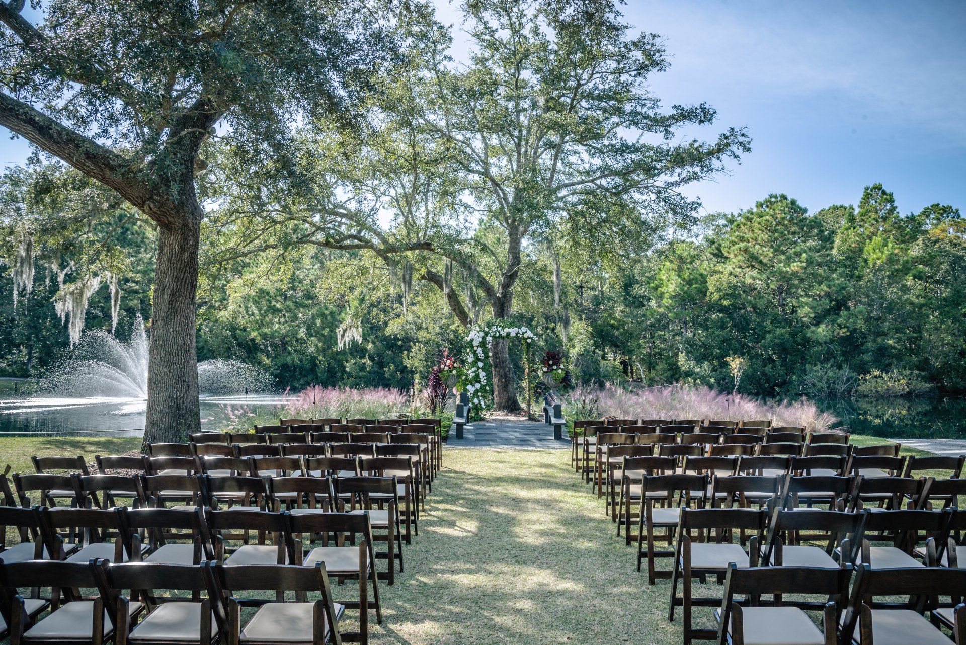 Outdoor wedding setup with rows of chairs facing an aisle under trees in a grassy garden.