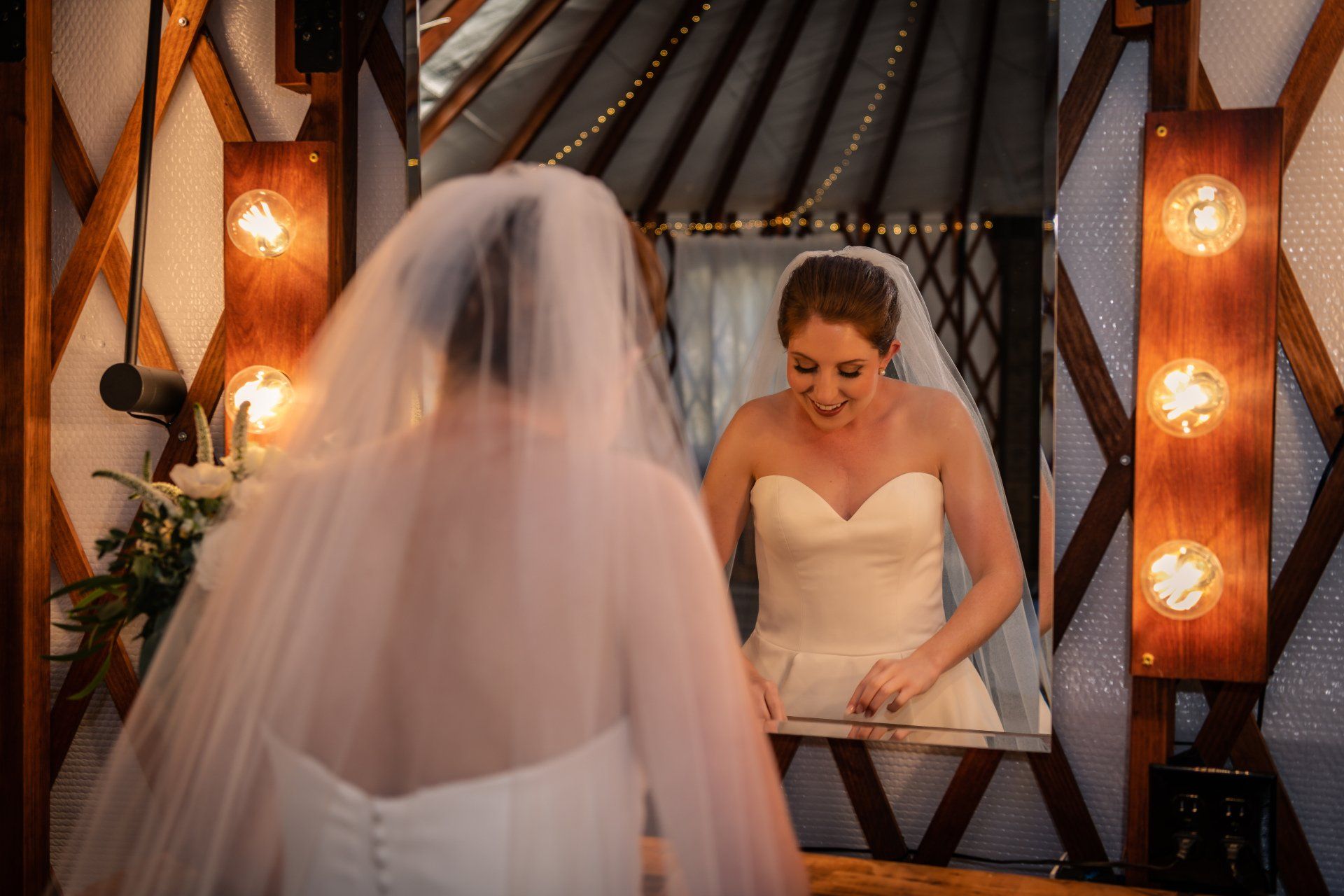 Bride in white dress and veil faces herself in a mirror, smiling in a warmly lit dressing room
