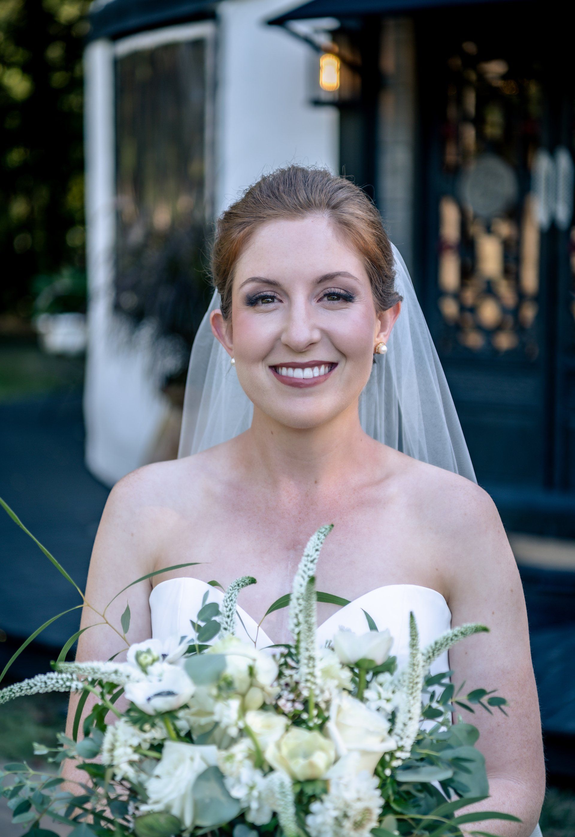 Smiling bride in a white dress and veil holding a white bouquet outdoors