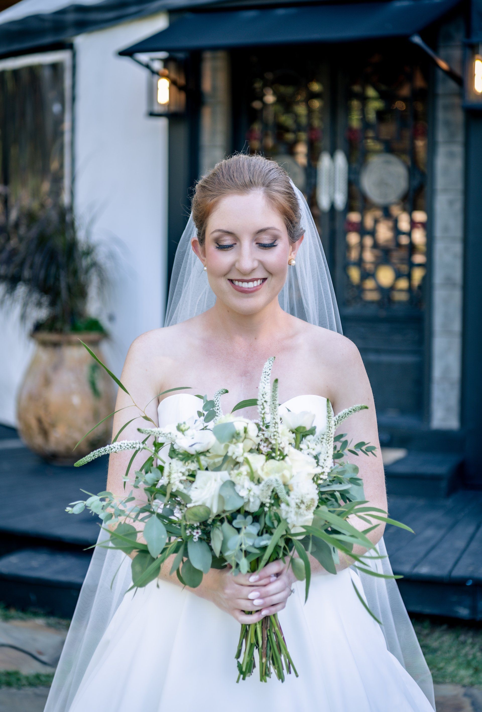 Bride in a white gown holding a bouquet of white flowers, standing outside a building and smiling.