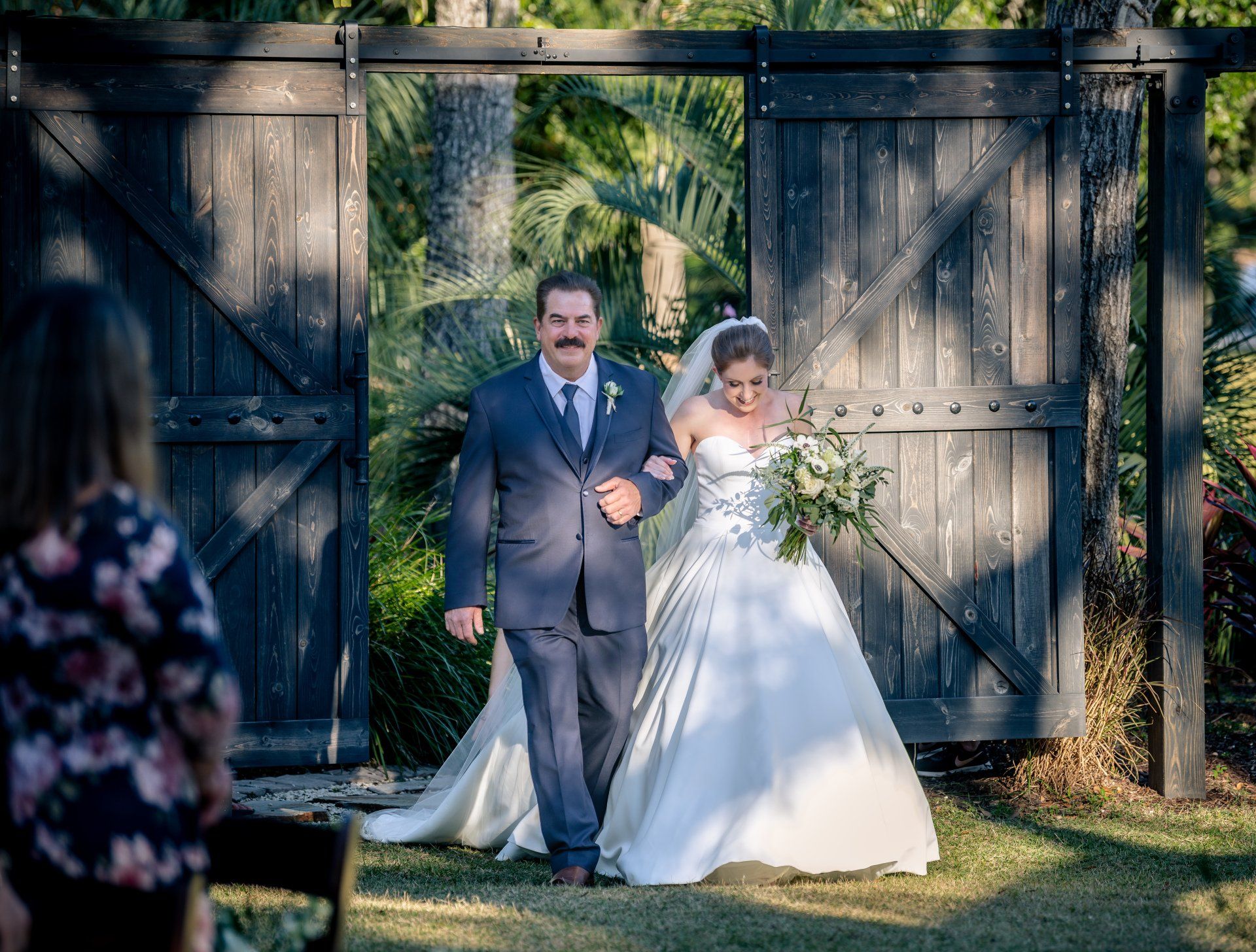 Bride and groom in navy suit and white gown walking through wooden gate in a garden