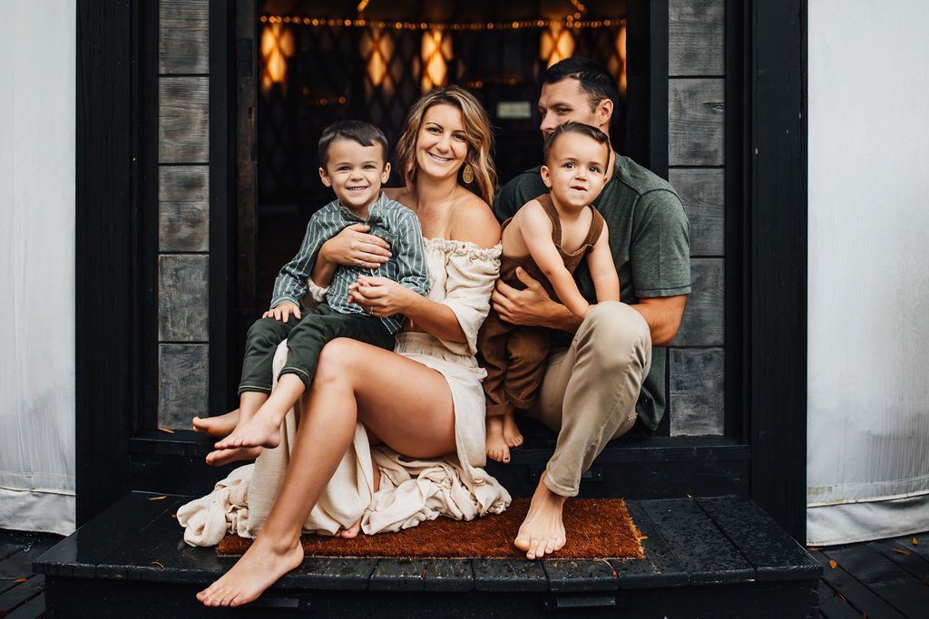 Family seated on a porch step in front of a warm-lit doorway, smiling and cuddling indoors.