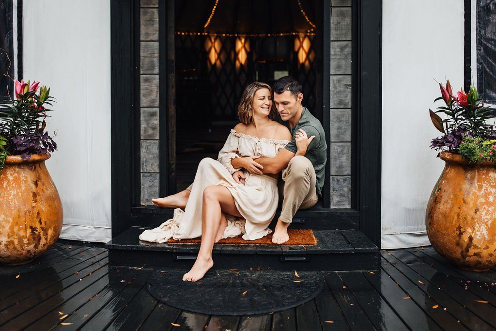 Couple sitting on a doorstep with flowers in rustic pots, warmly lit doorway behind them.