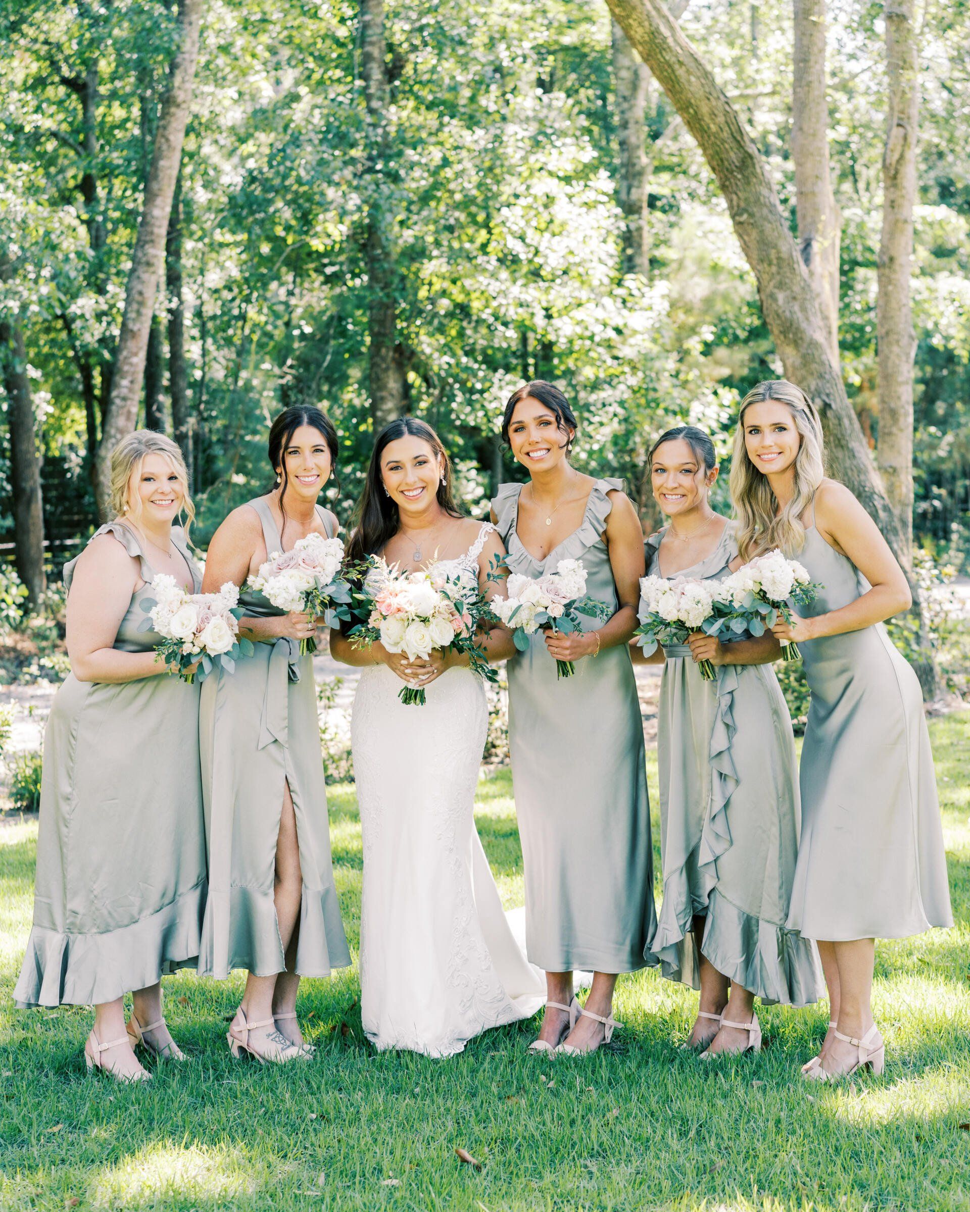 Five women in light dresses holding bouquets in a sunlit forest setting