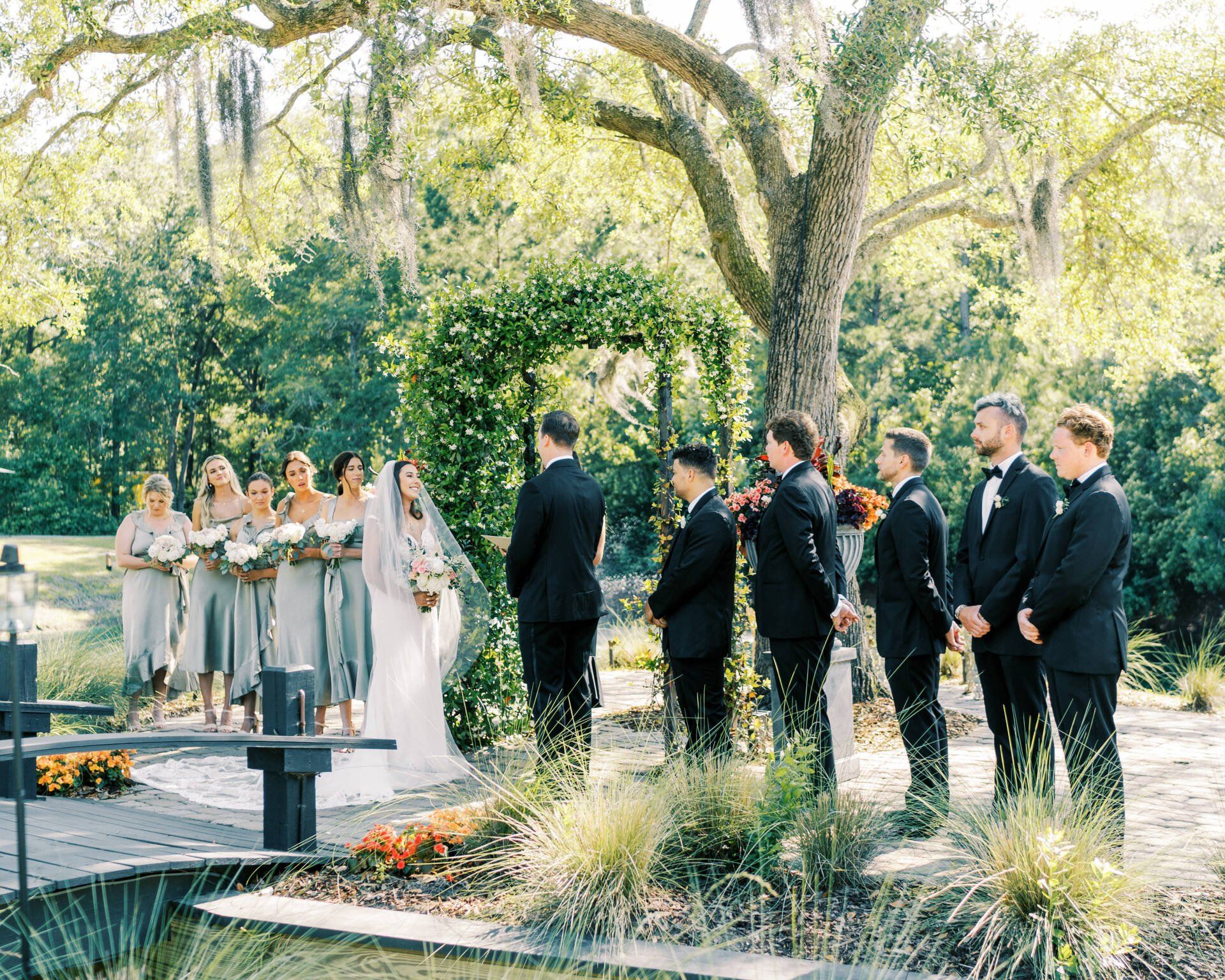 Outdoor wedding ceremony under trees, with bride and groom facing guests in formal attire.