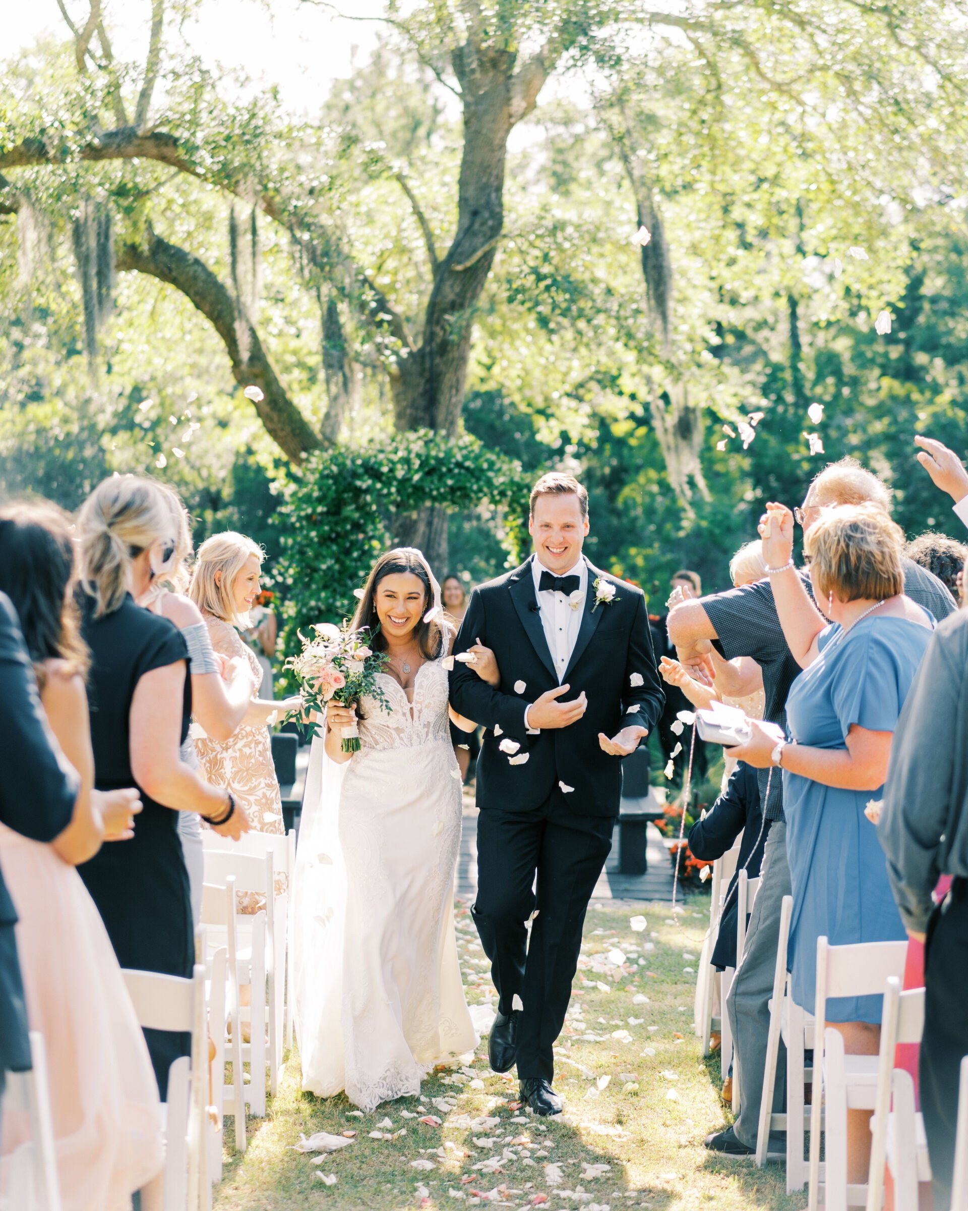 Wedding couple walking down an outdoor aisle, guests applauding beneath trees.