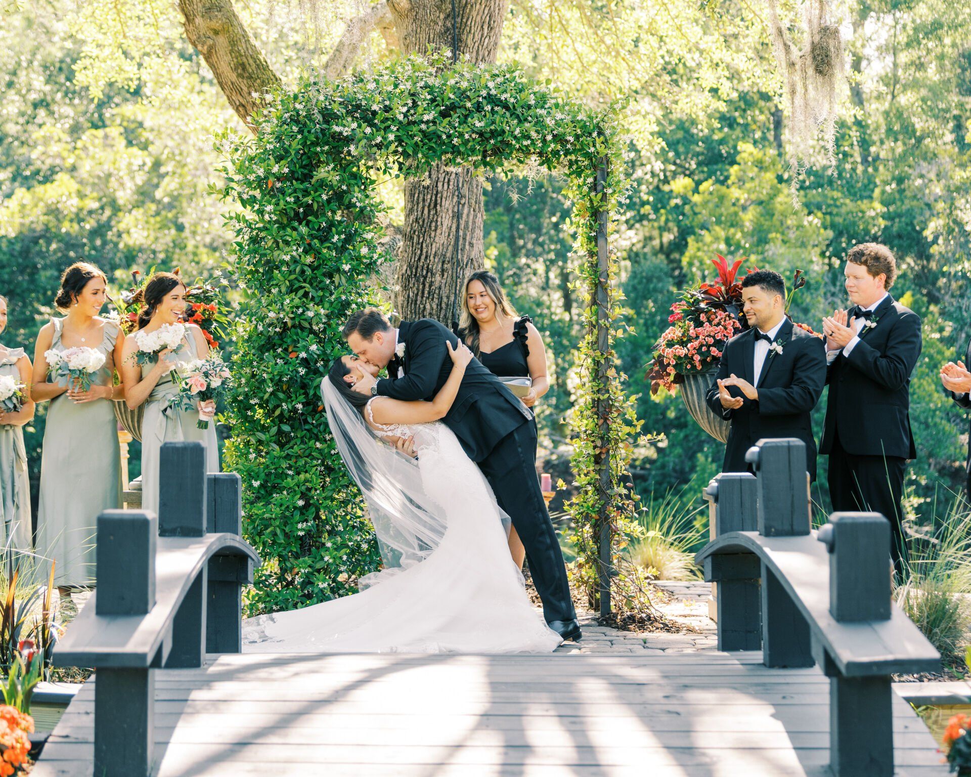 Bride and groom kiss at an outdoor wedding ceremony under a floral arch, guests applauding nearby.