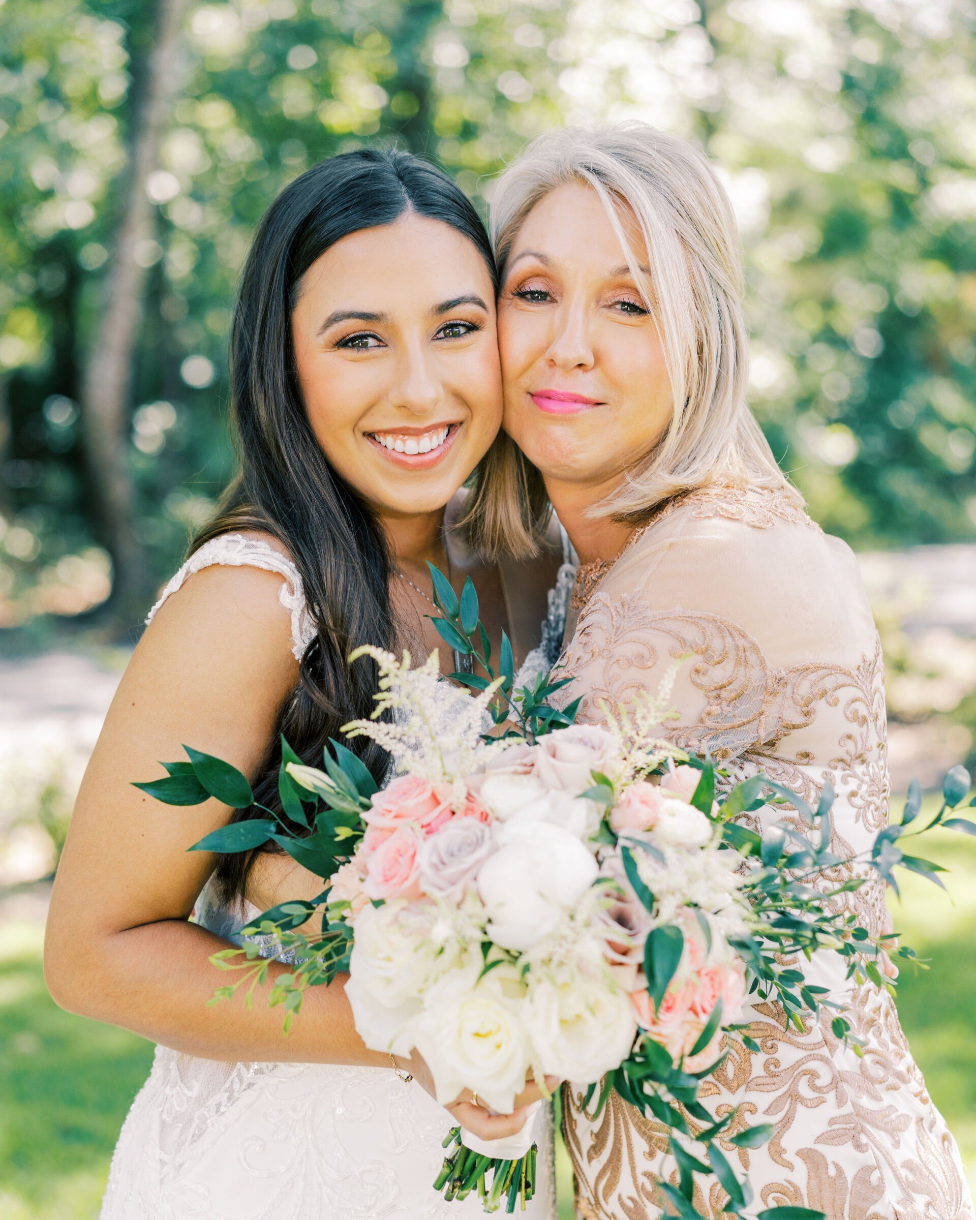 Two women in white dresses smiling and hugging, holding a bouquet outdoors in greenery.