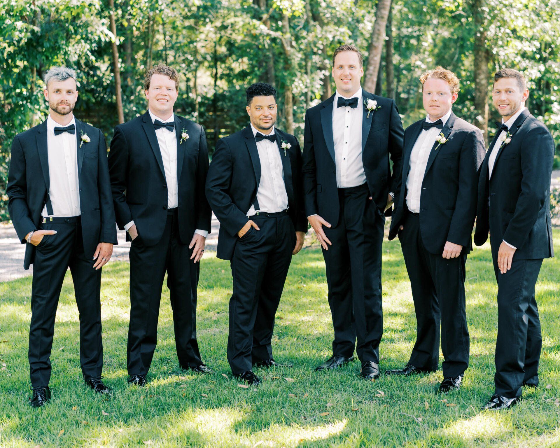 Six men in black tuxedos stand outdoors on grass with trees in the background.