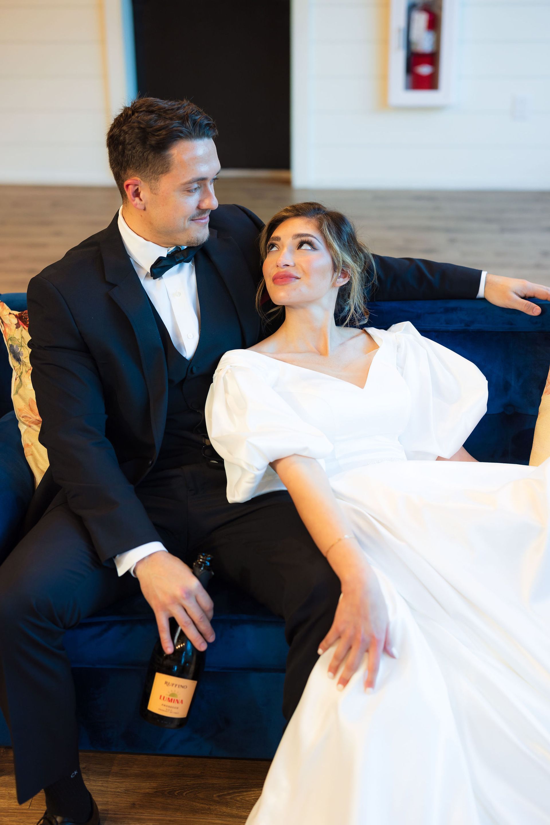 Couple in formalwear seated on a couch, smiling and leaning close, with a bottle in hand.