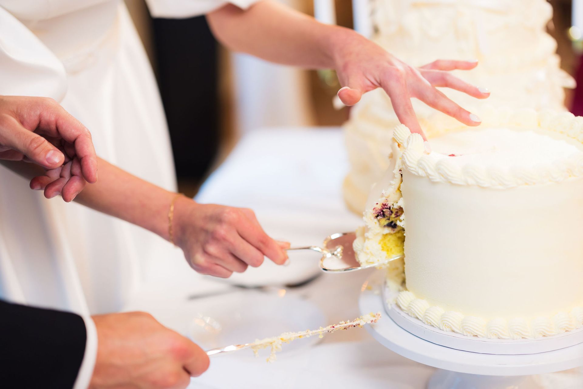 Hands cutting a white frosted wedding cake with a knife and server at a reception