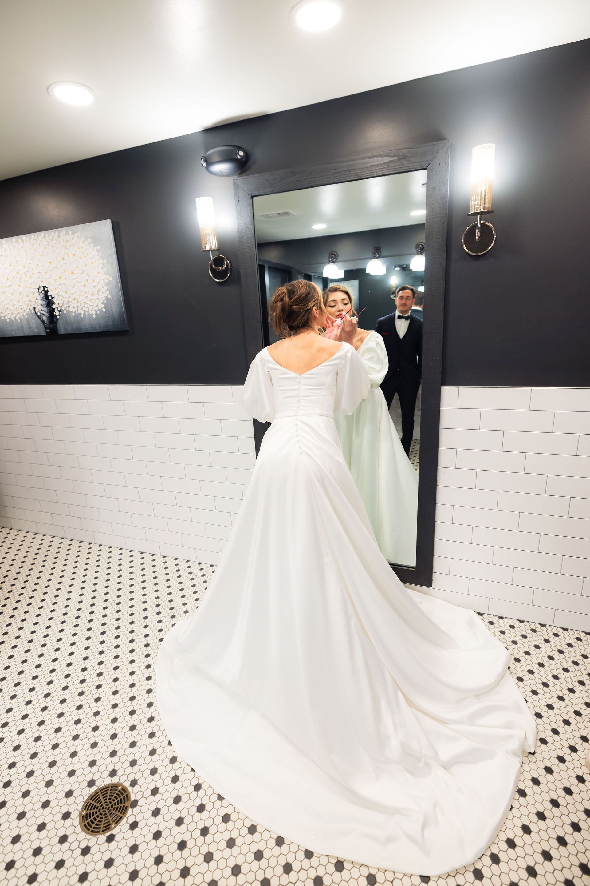 Bride in a white gown standing in a restroom, looking into a mirror while adjusting her dress.