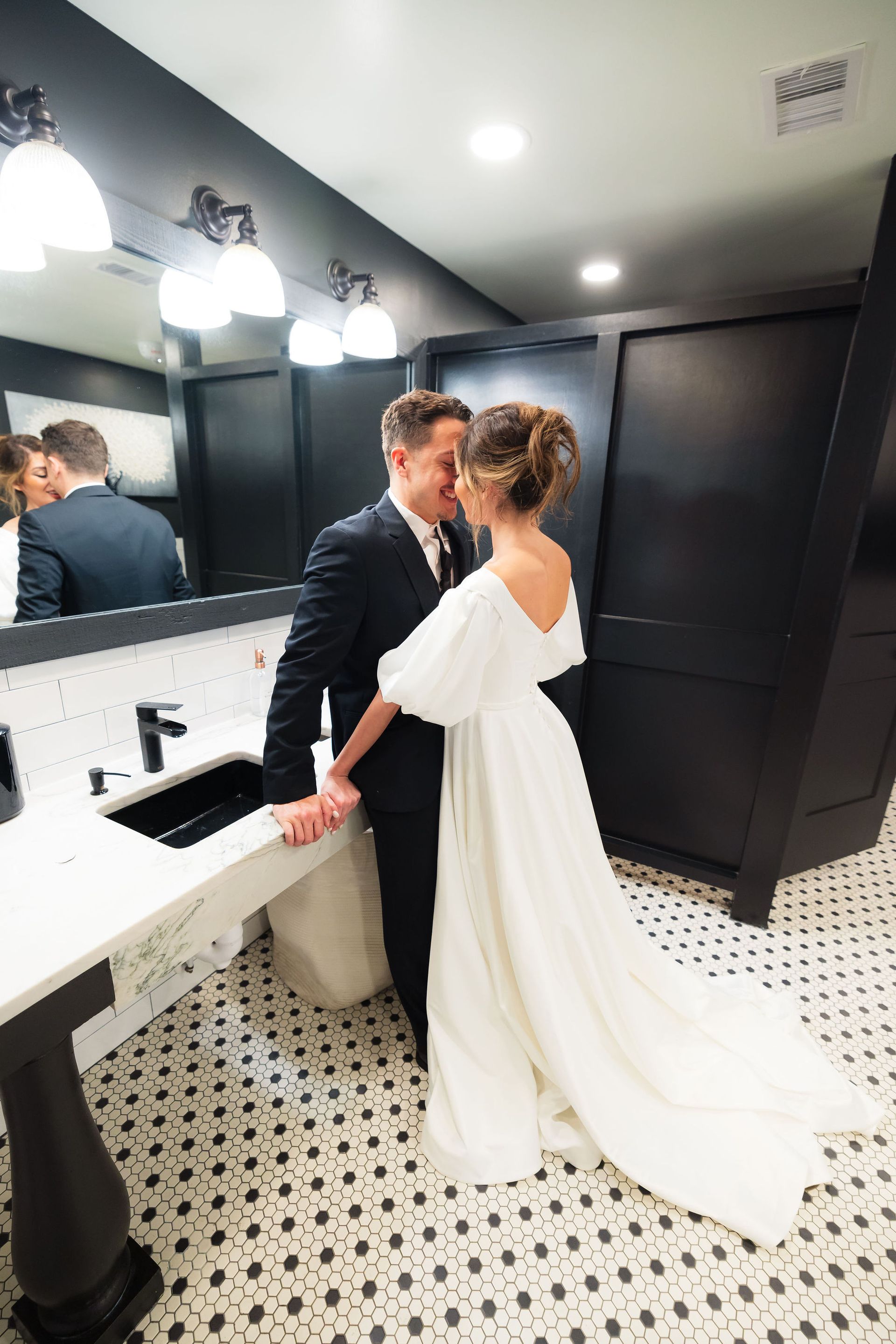 Bride and groom kissing in a modern black-and-white restroom, reflected in the mirror.