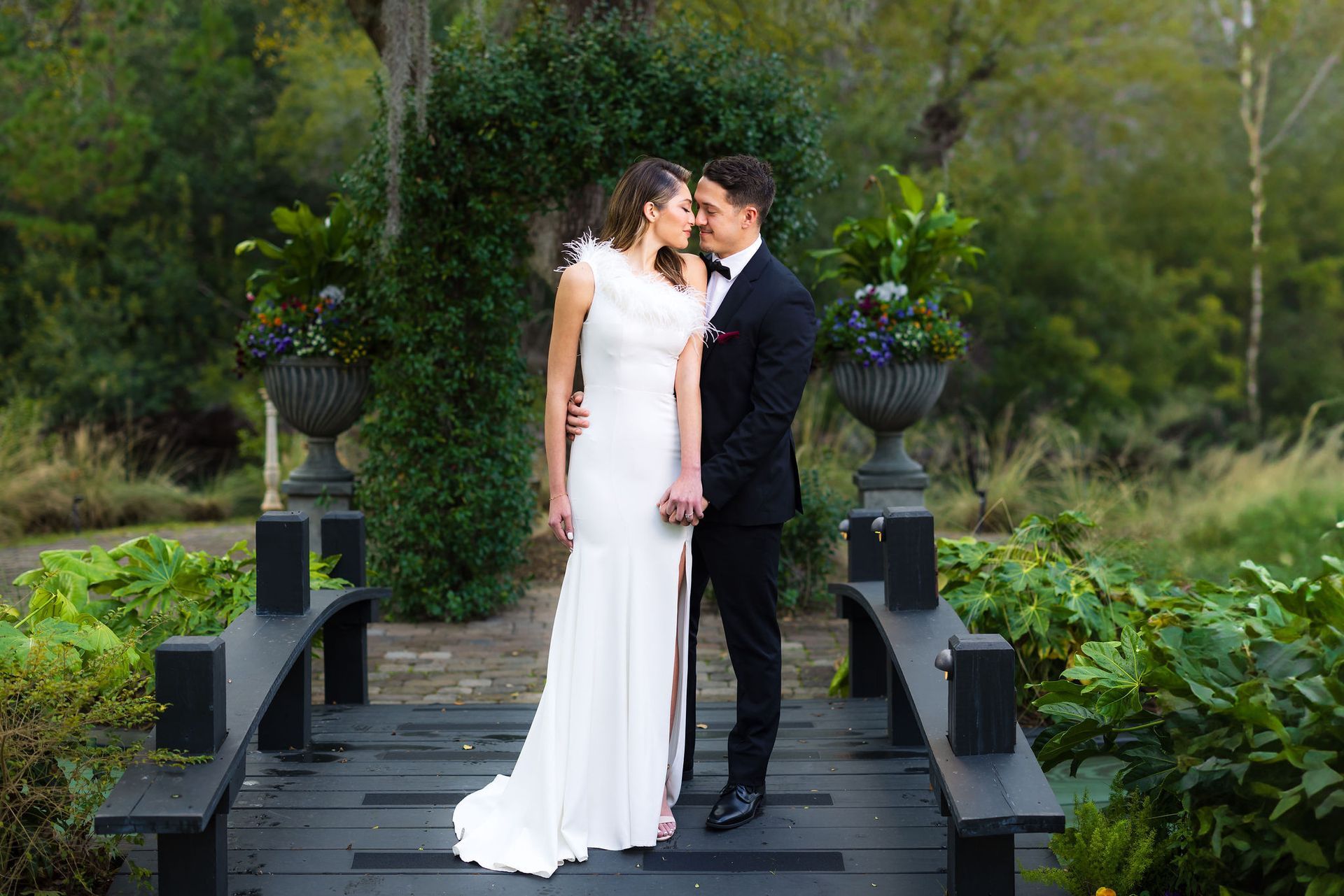 Bride and groom embrace on a dark wooden bridge surrounded by lush greenery.