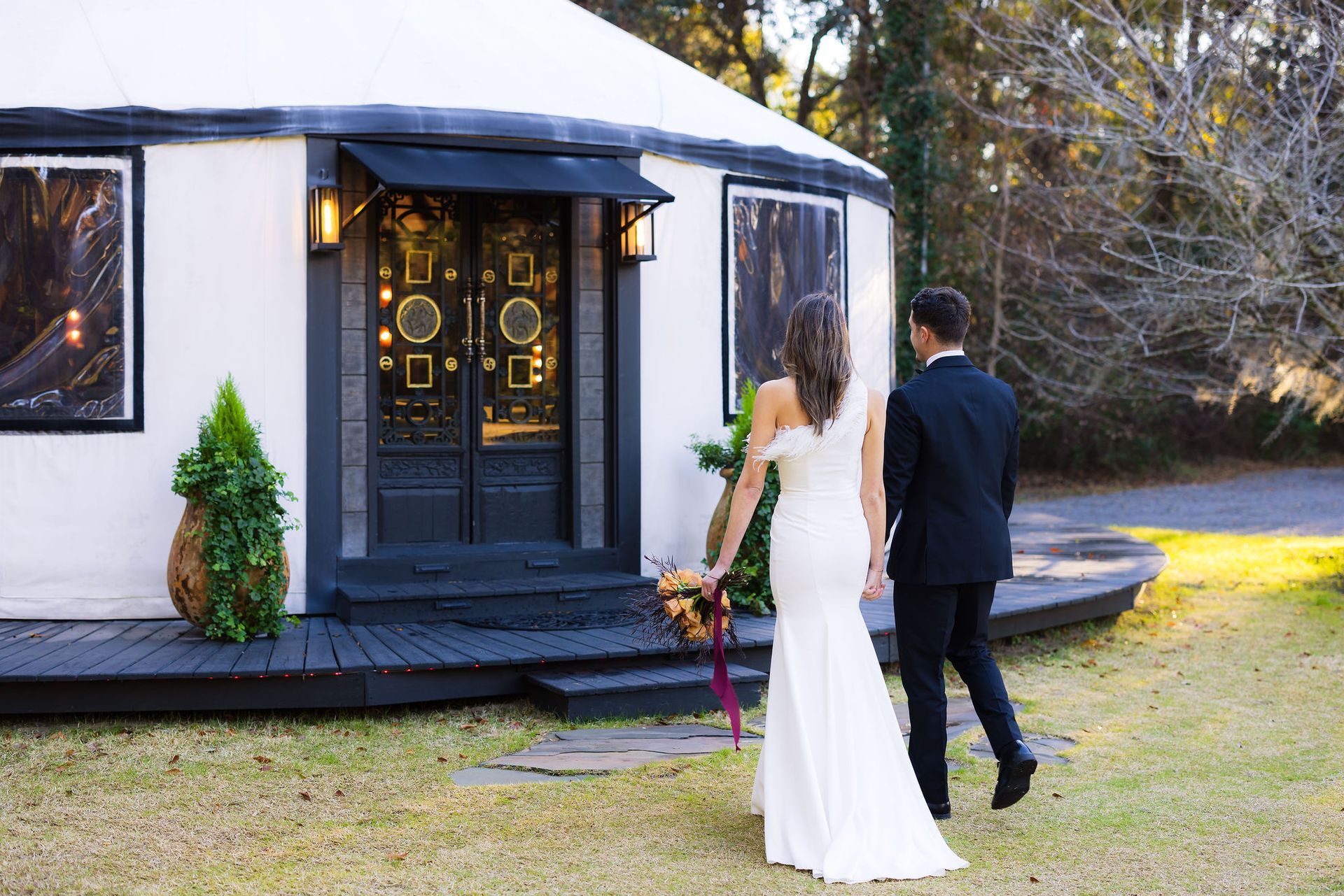 Bride and groom walking to a small white chapel in a grassy yard, seen from behind