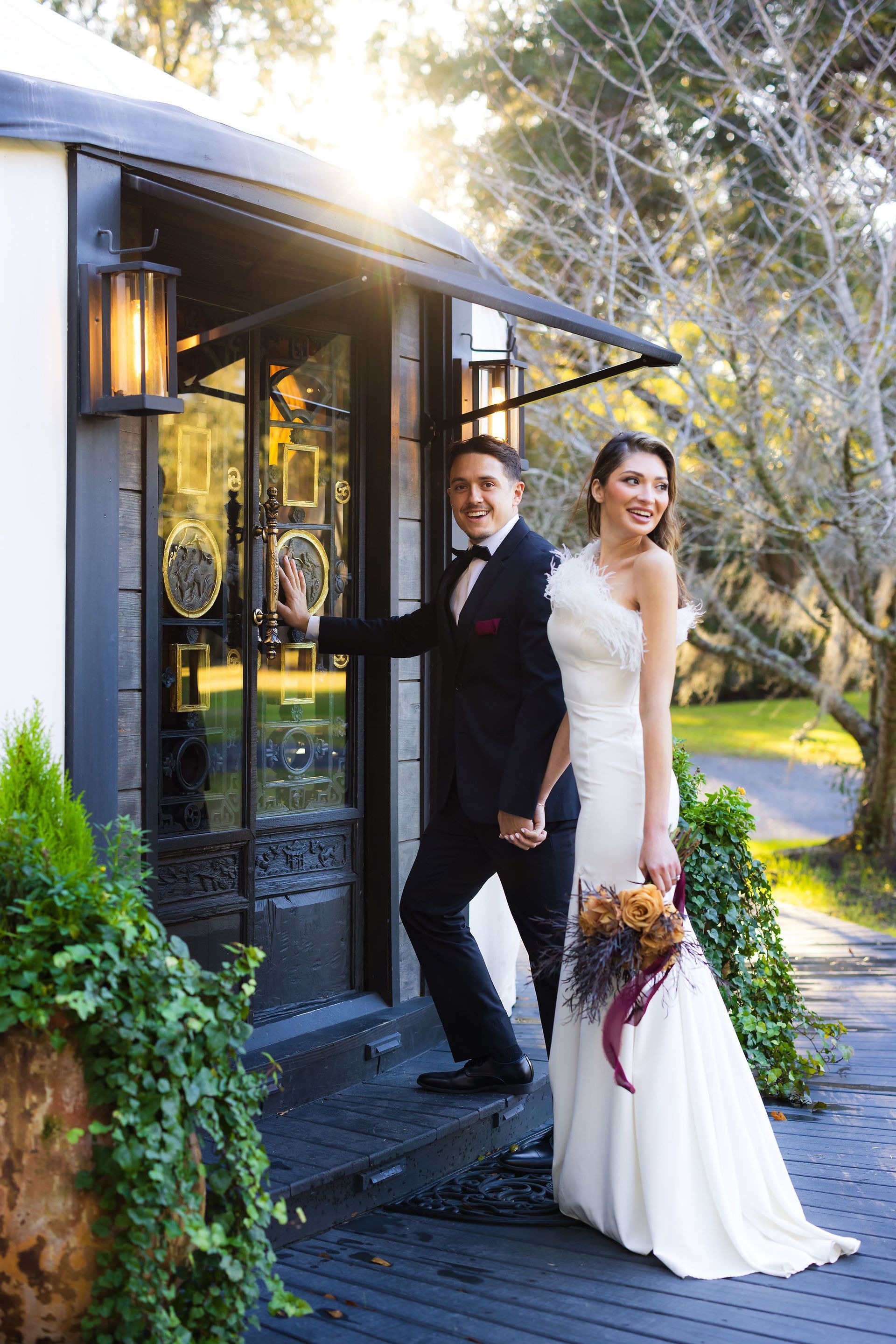 Bride and groom standing by a black doorway, smiling outdoors in wedding attire