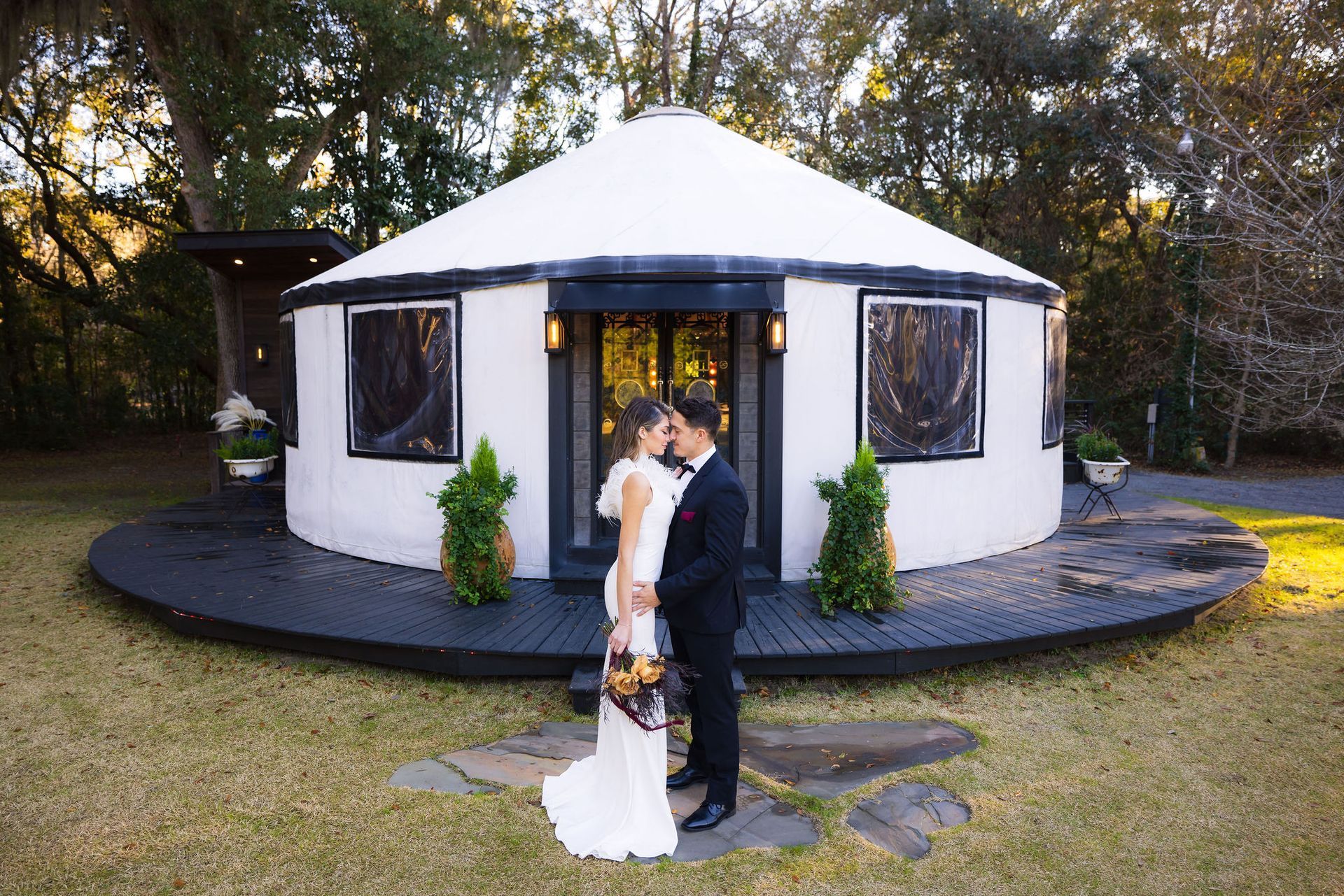 Bride and groom standing before a white round wedding pavilion with black trim, outdoors on grass